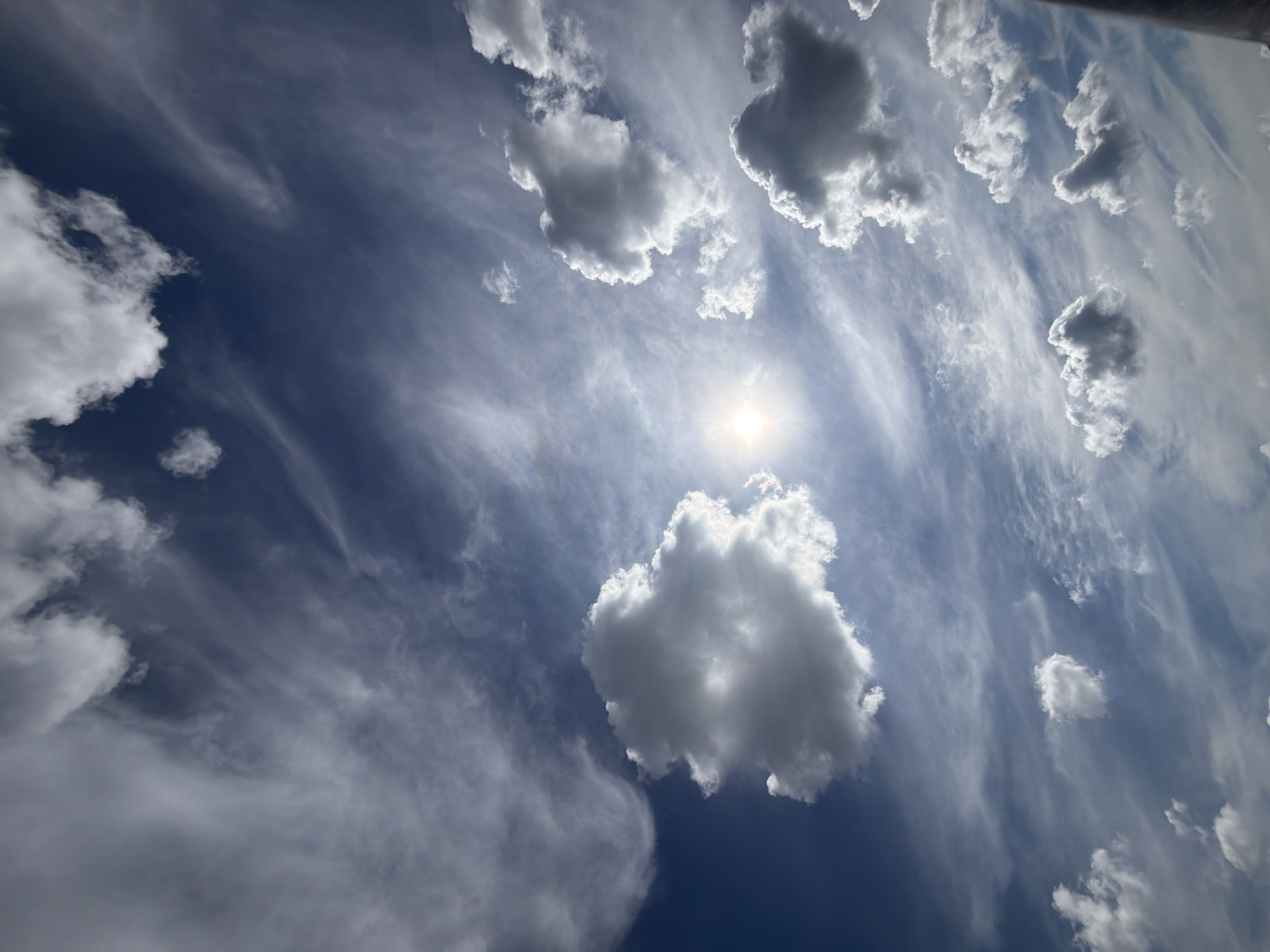 The image displays a vast expanse of the sky, predominantly filled with a mix of clouds against a deep blue backdrop. The sun is visible and partially obscured by clouds, casting a bright light. There are various types of clouds present, including wispy cirrus clouds and more dense cumulus clouds. The overall scene suggests an outdoor environment under a dynamic sky. The location of Findlay Township, United States of America, is not depicted or inferable from the image itself, as it solely focuses on the sky.