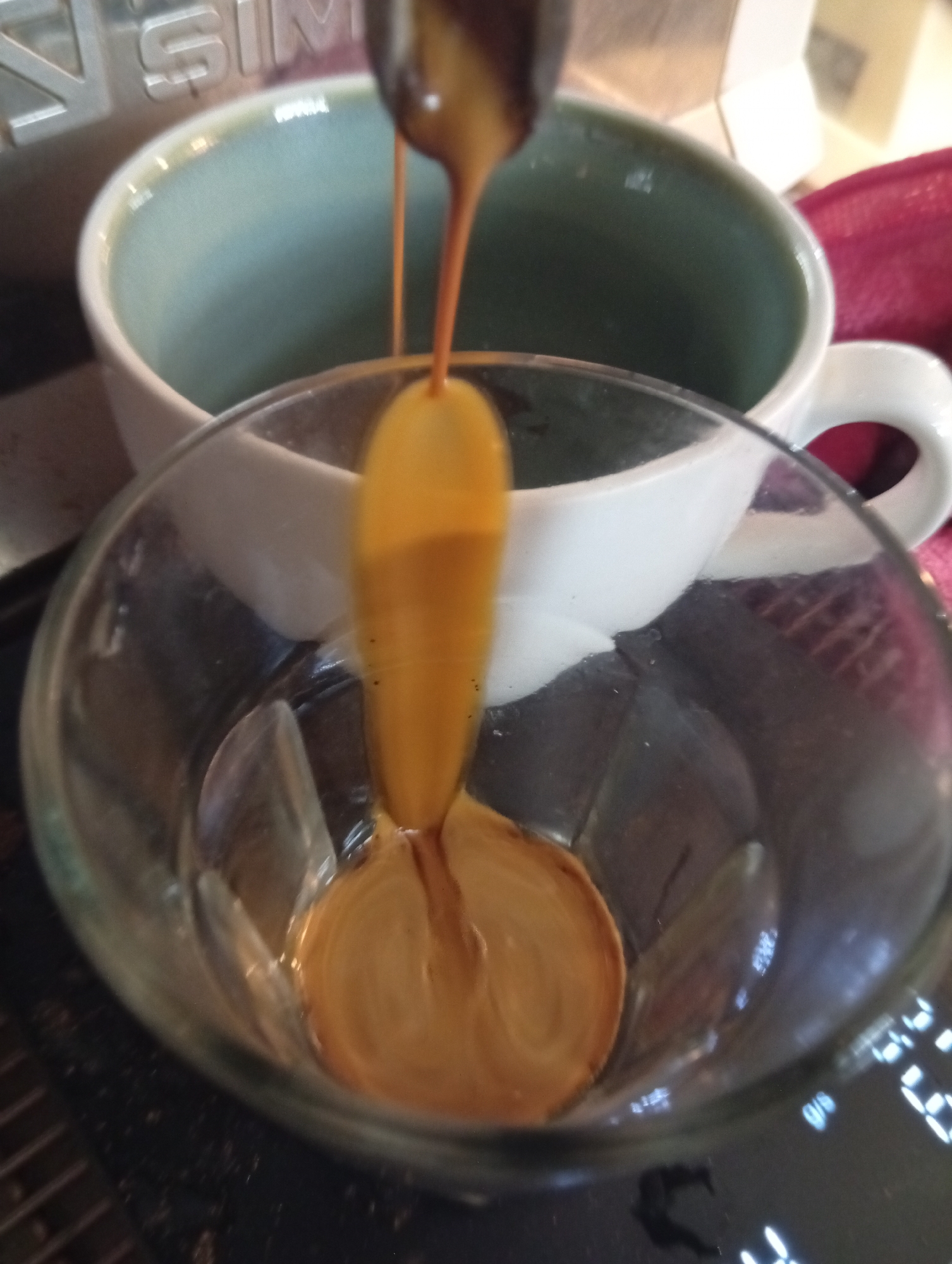 A close-up shows a clear glass with a thick stream of amber liquid pouring into it. The liquid is pooling at the bottom, forming a circular pattern. Behind the glass is a light blue mug. A coffee machine is partially visible in the background. A digital display is visible. The location and city cannot be determined from the image.
