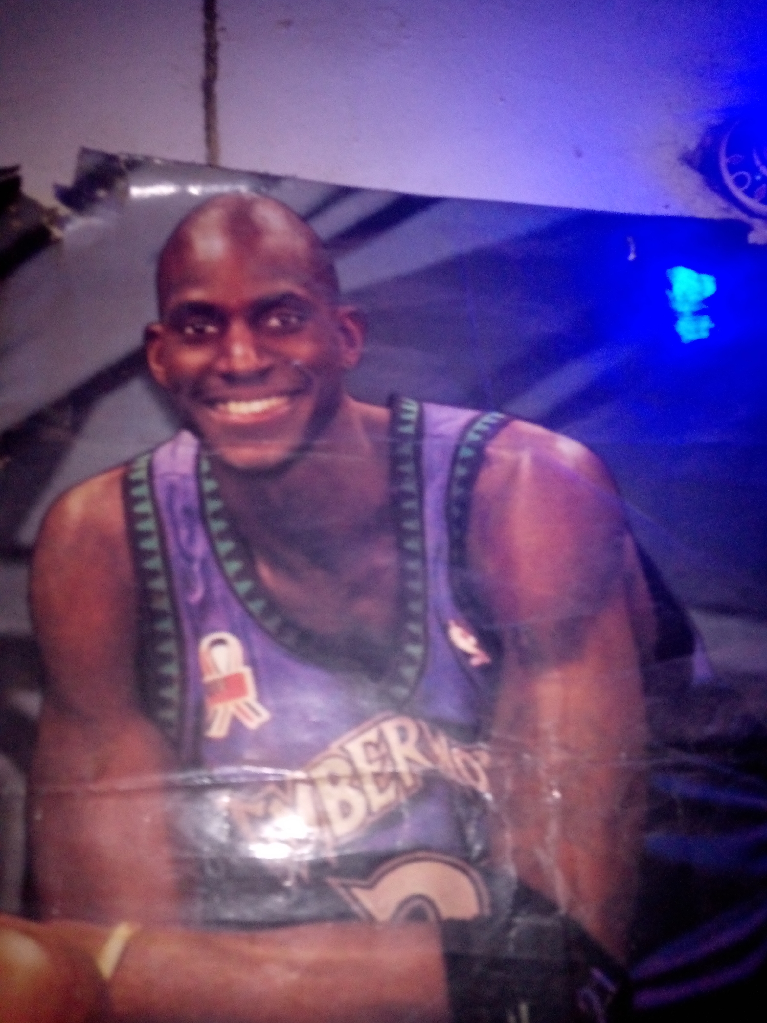 The image shows a close-up, slightly blurry photograph of a man wearing a purple basketball jersey. The jersey has a dark trim with a repeating triangular pattern and a logo with red and white elements on the left chest. The man is smiling broadly and has dark skin and a shaved head. The background is dark and out of focus, with some blue lighting visible on the right side, suggesting artificial lighting. There are no visible landmarks or elements that would specifically place this image in Jalingo, Nigeria. The image appears to be a poster or a photograph of a person, likely a basketball player.
