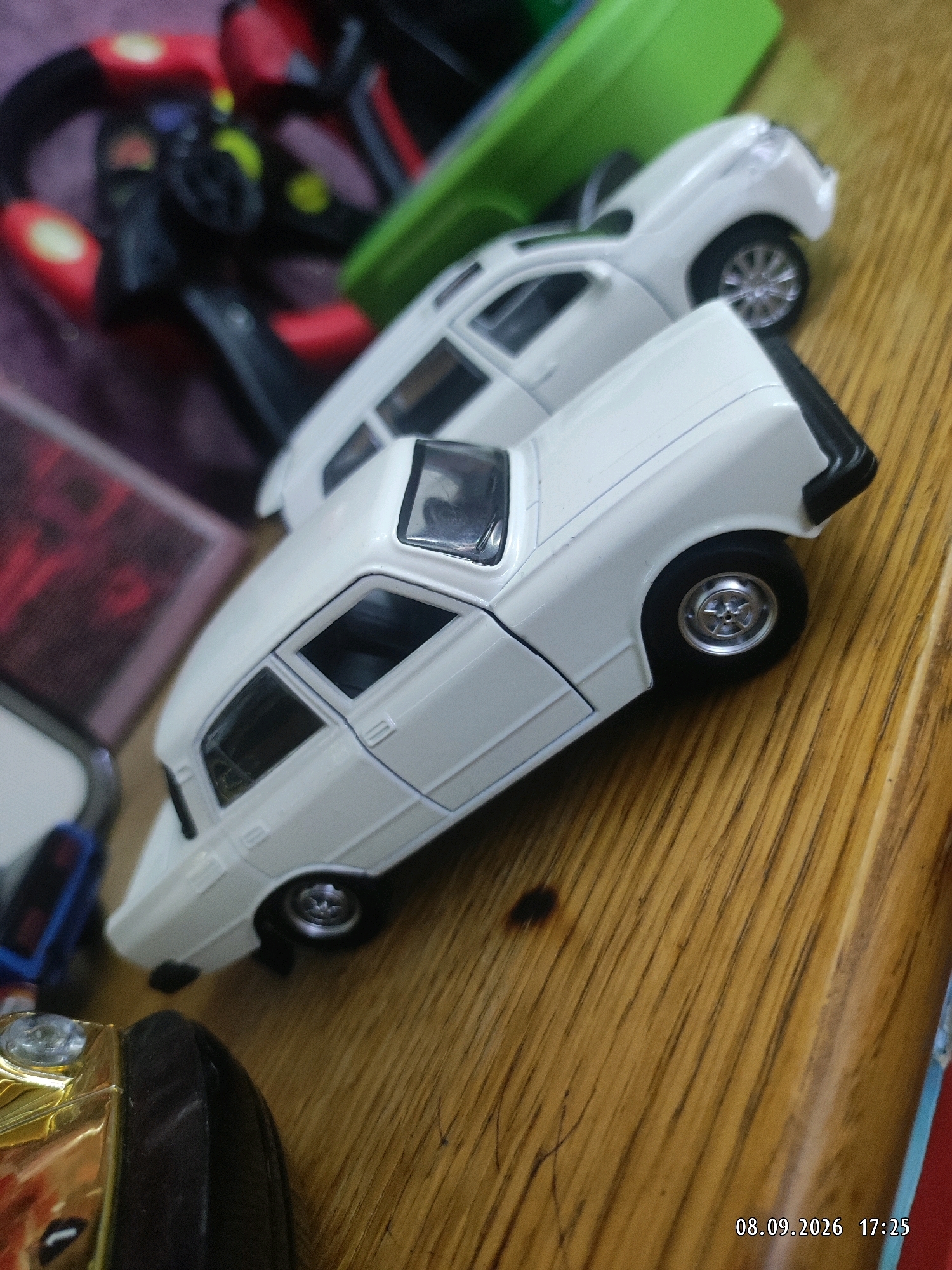 Two white toy cars are on a wooden surface. The car in the foreground is partially obscured by a gold and black object. The car in the background is a different model and appears to be a SUV. In the upper left corner, a blurry red and black object is visible. The environment appears to be indoors.