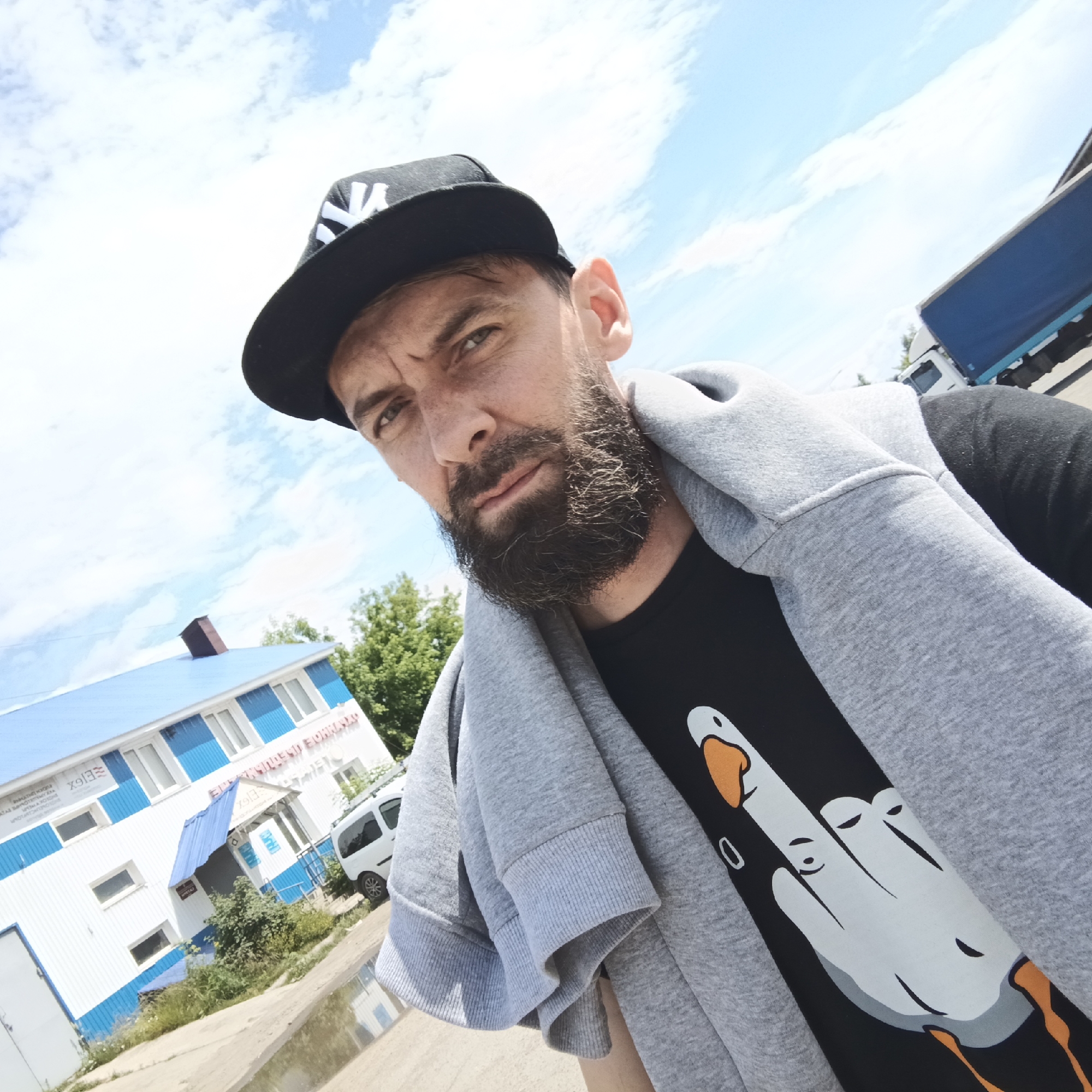 A man with a beard and a black baseball cap with a white logo is taking a selfie outdoors. He is wearing a black t-shirt with a graphic of a white duck-like figure with its middle finger extended, and a grey hooded sweatshirt draped over his shoulders. Behind him, a white building with a blue roof and blue trim is visible, with trees and a white van parked nearby. A blue truck is also partially visible in the background. The sky is cloudy and blue. The location is indicated as being in the city of Chaikovsky, Russia.