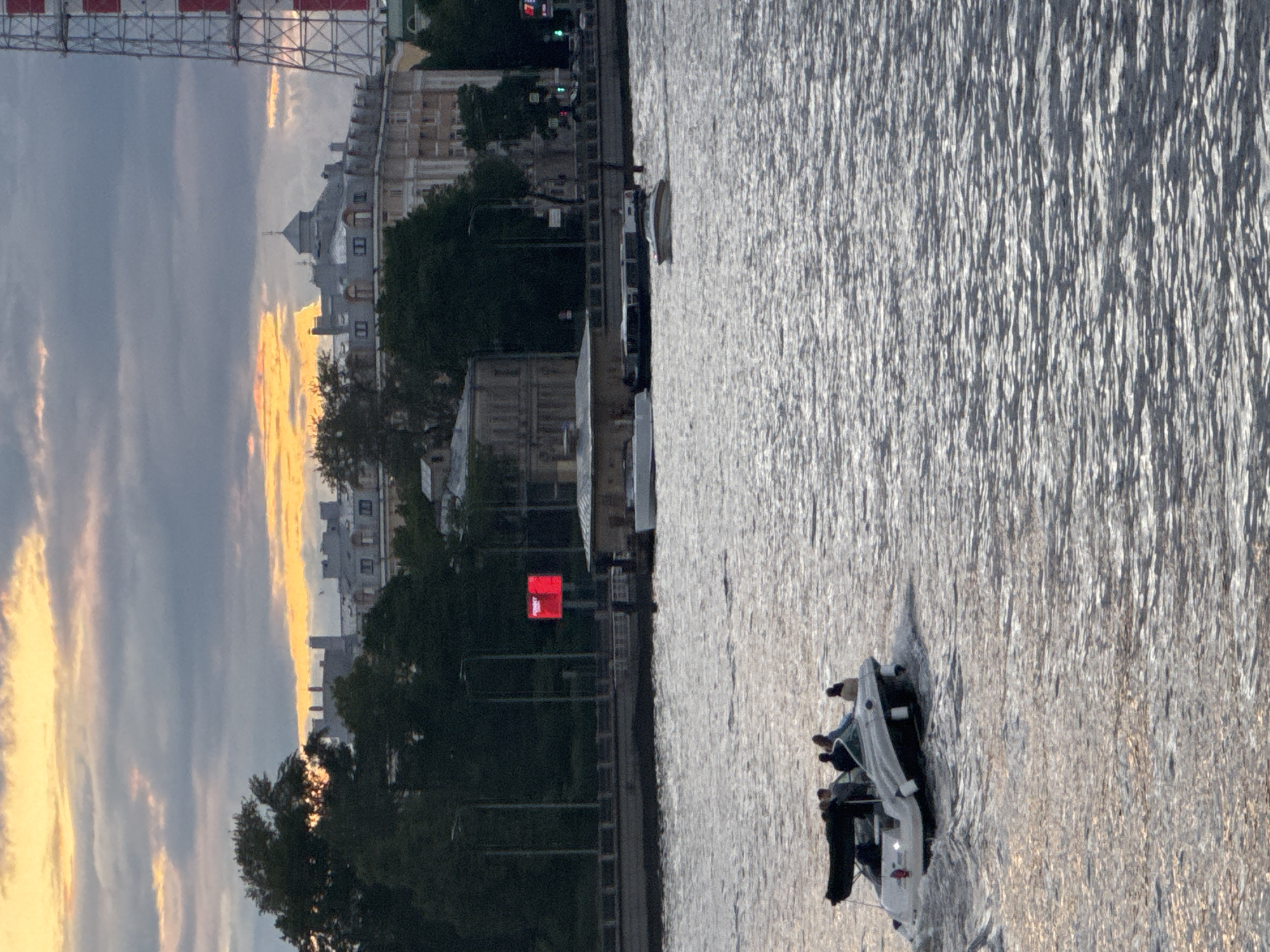A speed boat with five people on board is moving on the Neva River in Saint Petersburg, Russia. The boat is white with a black canopy and is creating a wake. In the background, several buildings and a tall industrial tower are visible. The sky is cloudy with patches of orange and yellow from the sunset.