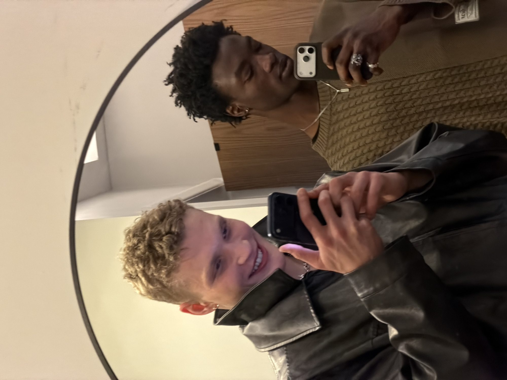 This is a mirror selfie featuring two young men in an indoor setting. The image is taken from a slightly tilted angle.
In the foreground, a man with light skin, short curly blonde hair, and an ear piercing is prominently featured. He is smiling widely, looking towards the camera reflected in the mirror, and holding a black smartphone with both hands, positioned to capture the image. He is wearing a black leather jacket over what appears to be a dark top.
Reflected behind him, and slightly to the right, is another man with dark skin and short, dark, curly hair. He is wearing a brown long-sleeved sweater with a distinctive textured, possibly cable-knit, pattern. A silver chain is visible around his neck, and he wears multiple rings on the fingers of his left hand, which is also holding a black smartphone. His expression is neutral as he looks towards the phone. A small, illegible tag is visible on the sleeve of his sweater.
The setting appears to be a room with plain light-colored walls and a background element that suggests wooden paneling or furniture. The mirror itself has a thin dark frame and reflects the subjects. The lighting is warm and artificial, common for indoor environments. The overall mood conveyed by the foreground subject's smile is cheerful. While the location context is London, United Kingdom, there are no specific visual cues within the image itself to confirm this.