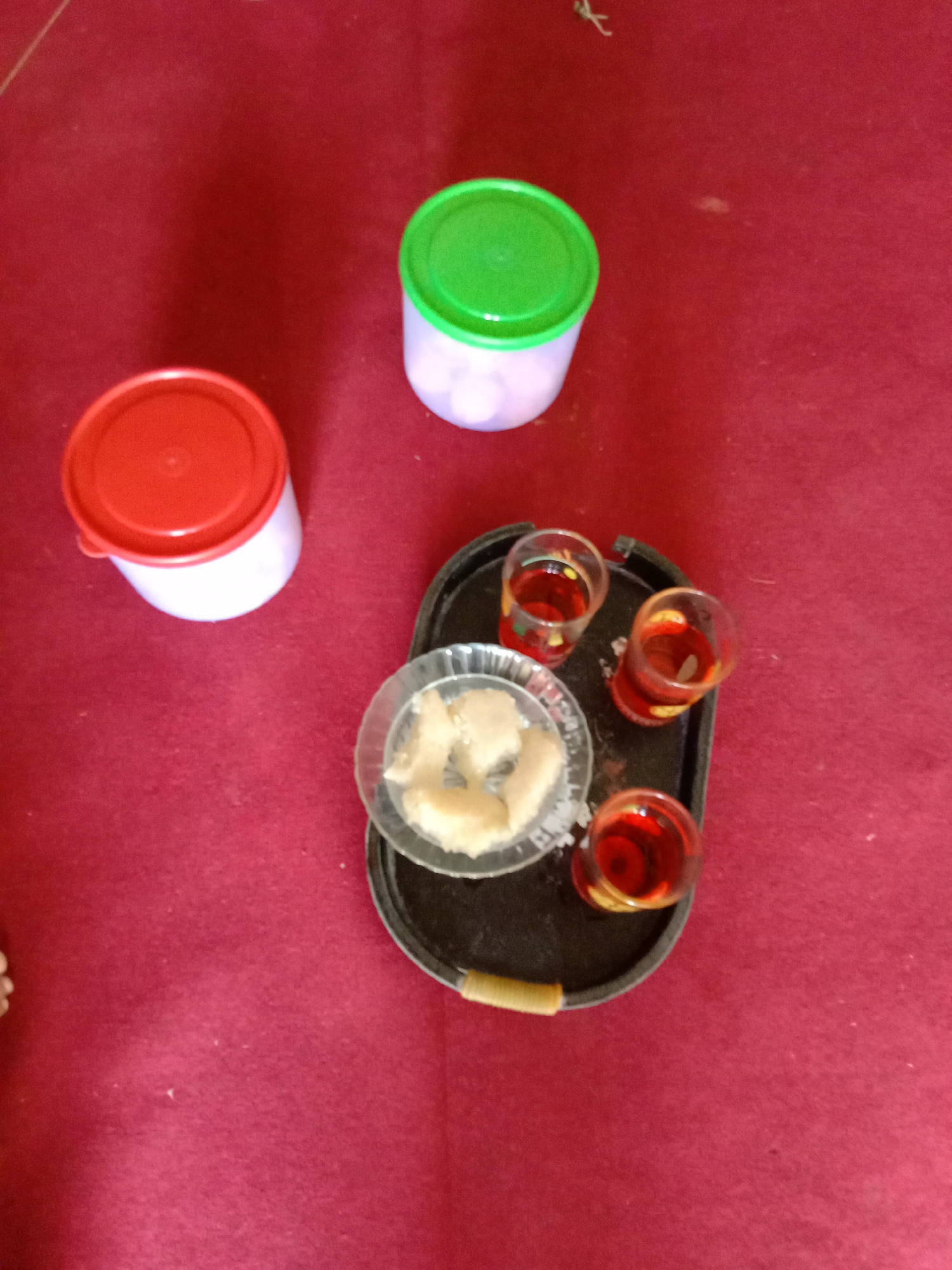 The image presents a still life scene on a dark red surface, likely a tablecloth. There are two circular plastic containers, one red-lidded and one green-lidded, positioned on the left and top of the frame, respectively. A dark-colored tray is placed centrally, containing three small glasses filled with a red liquid, presumably a beverage, and a glass dish holding light-colored, solid pieces, possibly food. The image appears to have been taken indoors. There is no information to confirm that this is in Blangkarieng, Indonesia.