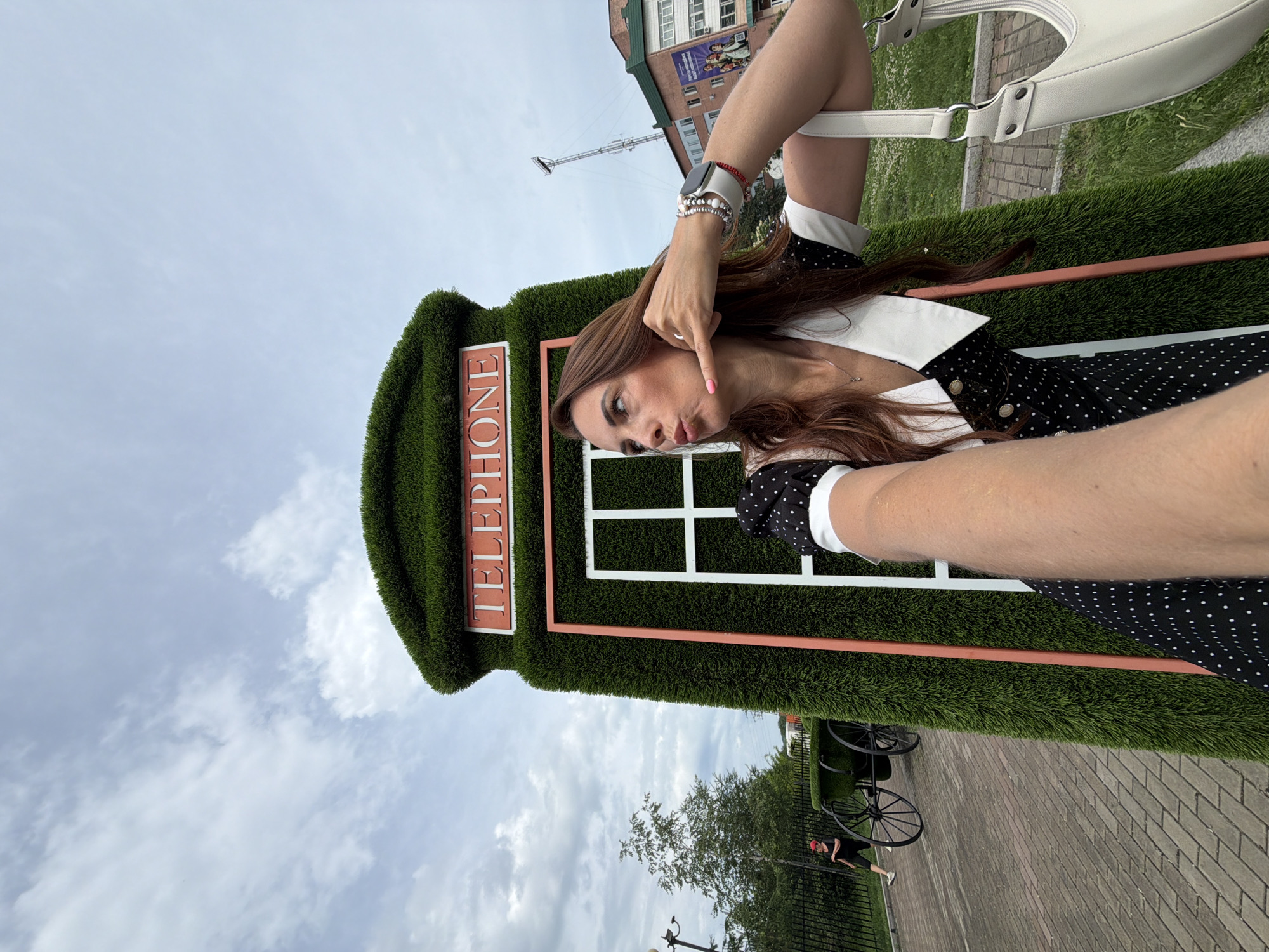 A woman with long brown hair is taking a selfie in front of a novelty, artificial grass-covered telephone booth. The booth is green and resembles a classic British red telephone box, but is covered in lush green artificial grass and has the word "TELEPHONE" in red letters at the top. The woman is wearing a black and white polka dot dress with a white collar and is making a "call me" gesture with her hand. A white handbag is slung over her shoulder. In the background, a person in a red hat is walking past a decorative carriage. Buildings and a transmission tower are visible in the distance under a cloudy sky. This scene is located in Birobidzhan, Russia.