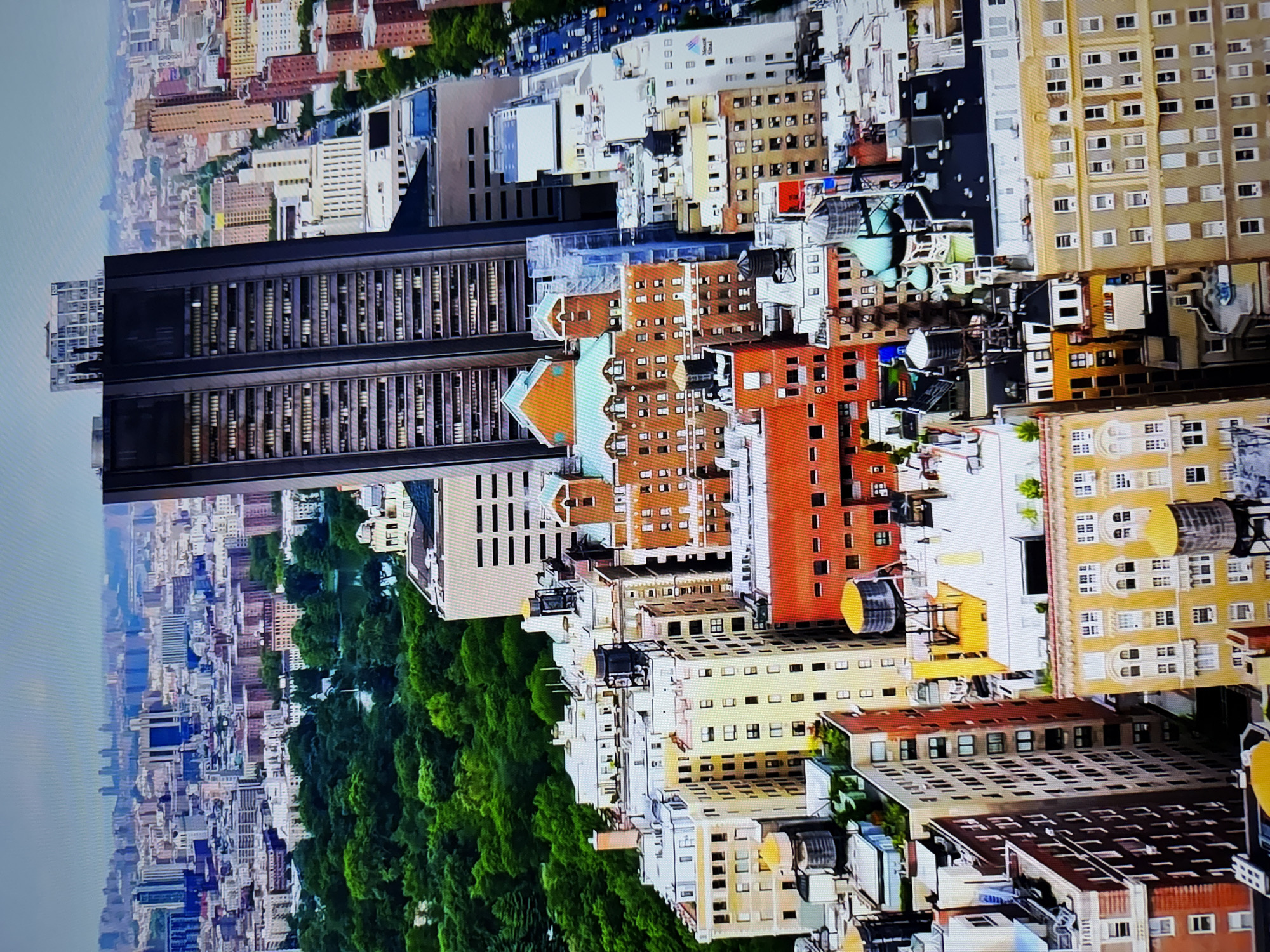 The image is an aerial view of a dense urban landscape, dominated by buildings of varying heights and architectural styles. In the foreground and midground, numerous apartment buildings with distinct brick facades and rooftop water towers are visible. Many of these structures feature repetitive window patterns, indicative of residential blocks. A prominent, dark, rectangular skyscraper stands out in the midground, its facade featuring rows of windows and a stepped top. Green trees are abundant, suggesting proximity to a park or densely treed area, particularly in the background where a large expanse of green foliage forms a distinct contrast to the urban development. Beyond this, a hazy cityscape with numerous smaller buildings stretches to the horizon under a pale sky. A street with traffic is visible on the right side of the image, running parallel to some of the buildings. The scene depicts a high-density urban environment, likely a major city, with a mix of older and more modern architecture. The location is identified as Burgas, Bulgaria.