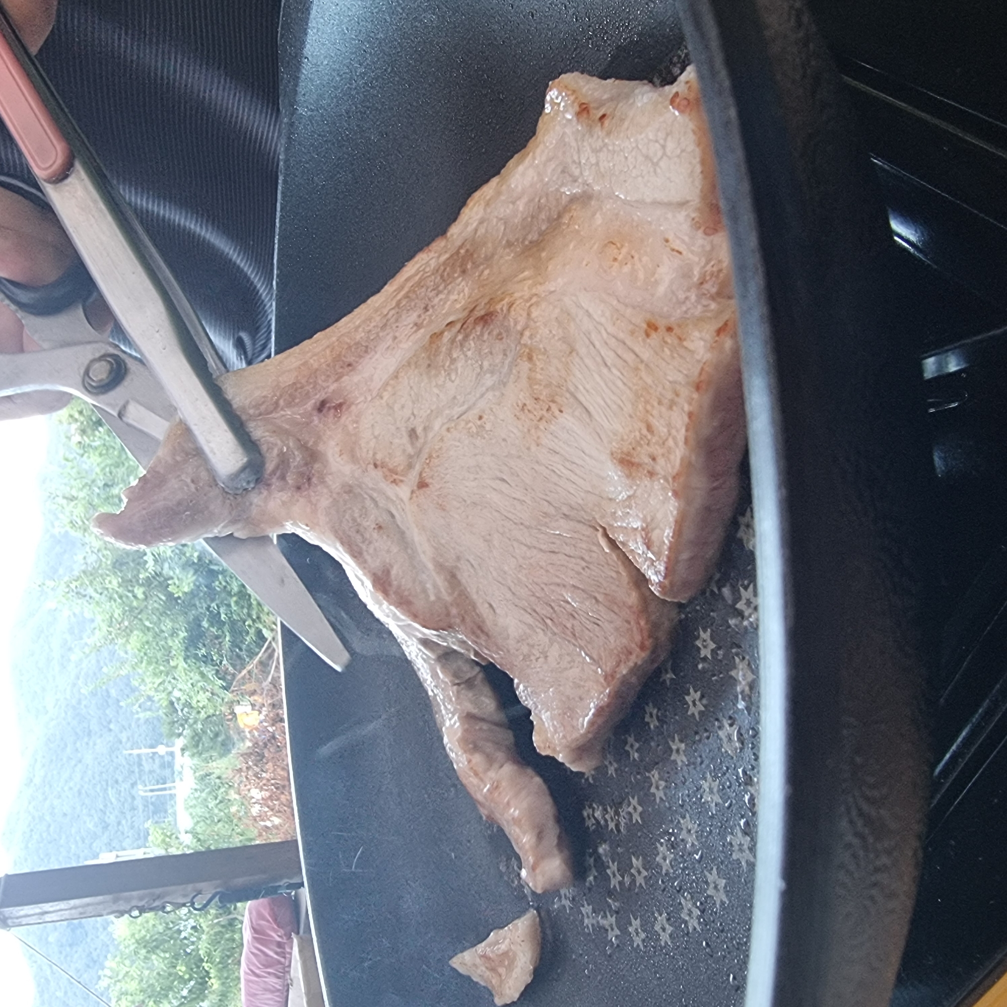 A pair of silver-handled scissors is being used to cut a piece of raw meat in a black frying pan. The meat is a pale pink color and appears to be a cut of pork. The frying pan is on a stovetop, and the background shows a blurry view of trees and mountains, suggesting an outdoor setting, possibly in Dunnae-myeon, South Korea. There is also a red cushion visible in the background.