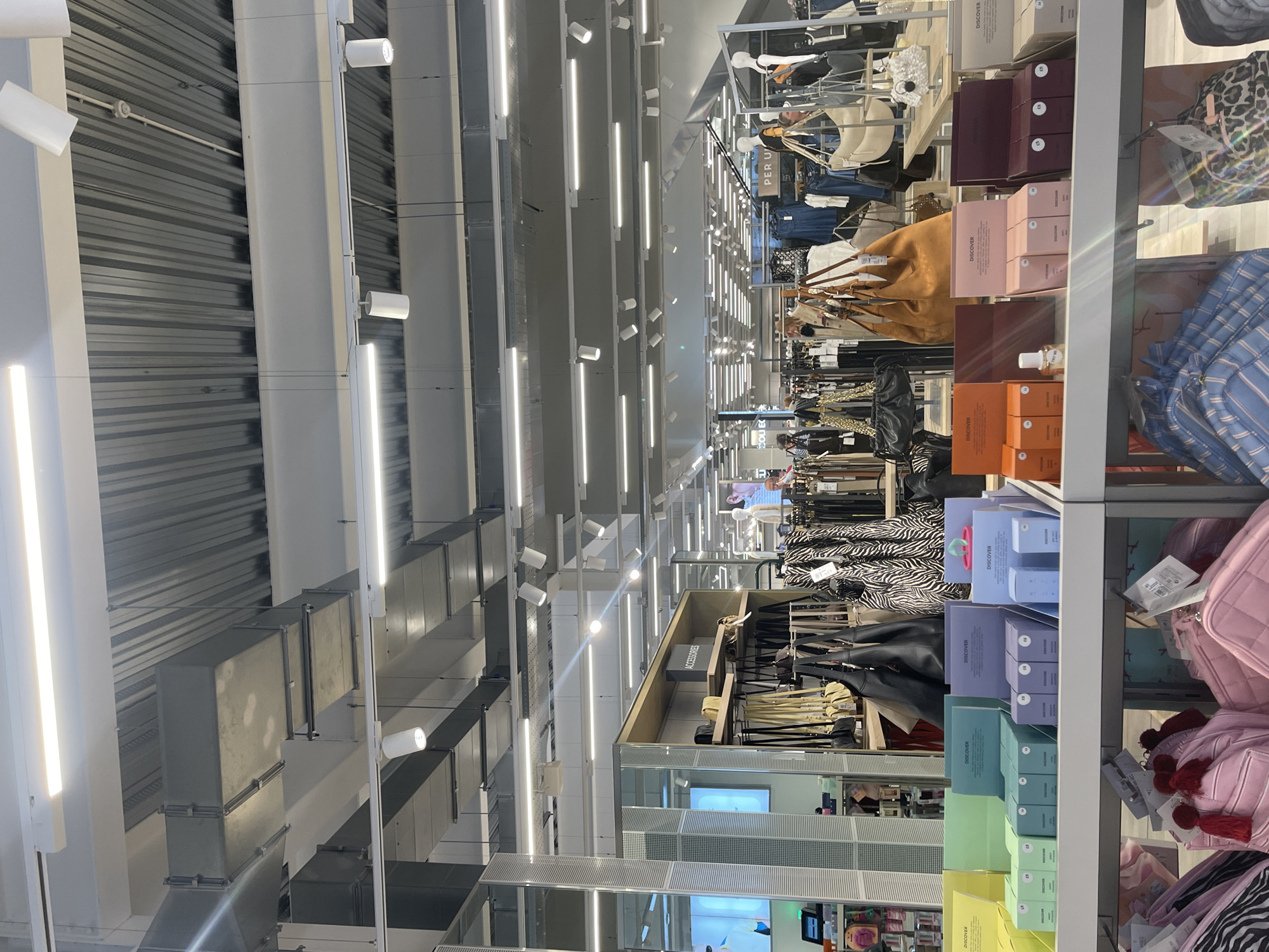 The image displays the interior of a brightly lit, modern retail store, likely a fashion or department store, situated in Maidstone, United Kingdom. The perspective is taken from within an aisle, looking across various merchandise displays towards the rear of the store.
The ceiling features an industrial design with exposed corrugated metal panels and prominent rectangular air ducts. A comprehensive lighting system is integrated into the ceiling, consisting of long, linear fluorescent light fixtures providing general illumination, alongside numerous smaller, circular track lights directed at display areas. The ceiling and structural elements are predominantly white and light grey.
On the right side of the frame, multiple retail display units showcase a wide array of products. These include shelves stacked with boxed items, presented in a spectrum of pastel colors such as maroon, pink, orange, light blue, lavender, light green, and yellow. Many of these boxes are labeled "DISCOVER" and some have visible price tags indicating "£3" or "£5". Below these boxed items, various handbags are displayed, including a large tan hobo bag, a leopard print bag, a striped blue and white bag, and several pink quilted bags with decorative tassels.
Further into the background, clothing racks hold different garments, including denim and items with black and white zebra print. A sign reading "ACCESSORIES" is visible above a display, and another partially obscured sign bears the text "PER U". Mannequins are faintly discernible in the distance, modeling clothing. The store appears organized and well-maintained, suggesting it is open for business during regular operating hours. No direct human activity is captured, but the setup implies an environment for shopping and browsing.