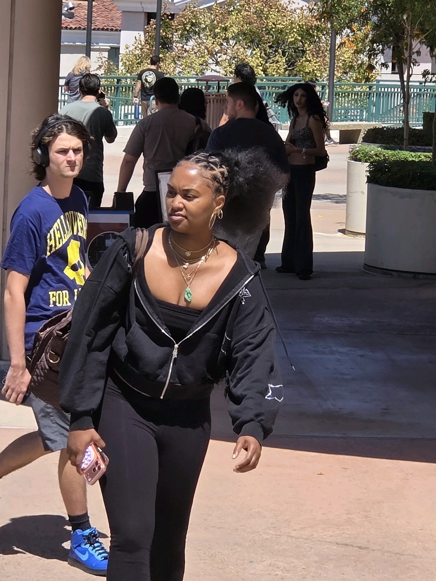 In Glendale, California, on a sunny day, two individuals are captured in a street-level view. In the foreground, a young Black woman with braided hair pulled back walks towards the right of the frame, wearing a black zip-up hoodie, a black strapless top, and black leggings. She is adorned with several necklaces, including a prominent green pendant, and hoop earrings. She holds a rose gold smartphone in her right hand. To her left, a young White man with medium-length brown hair and headphones on his ears walks in the same direction, wearing a royal blue t-shirt with yellow lettering that reads "HELLUVA FOR..." and gray shorts. He has a brown messenger bag slung over his left shoulder.
In the mid-ground, a White woman with long, dark, curly hair is visible walking away from the camera, wearing a leopard-print tank top and dark pants, with a black backpack. Behind her, several other individuals are present, some walking and some standing. The environment appears to be an outdoor walkway or plaza, with buildings and trees visible in the background. The walkway is paved with a reddish-brown material, and there are large, cylindrical planters with greenery. The lighting suggests it is daytime, with strong shadows cast on the ground.
