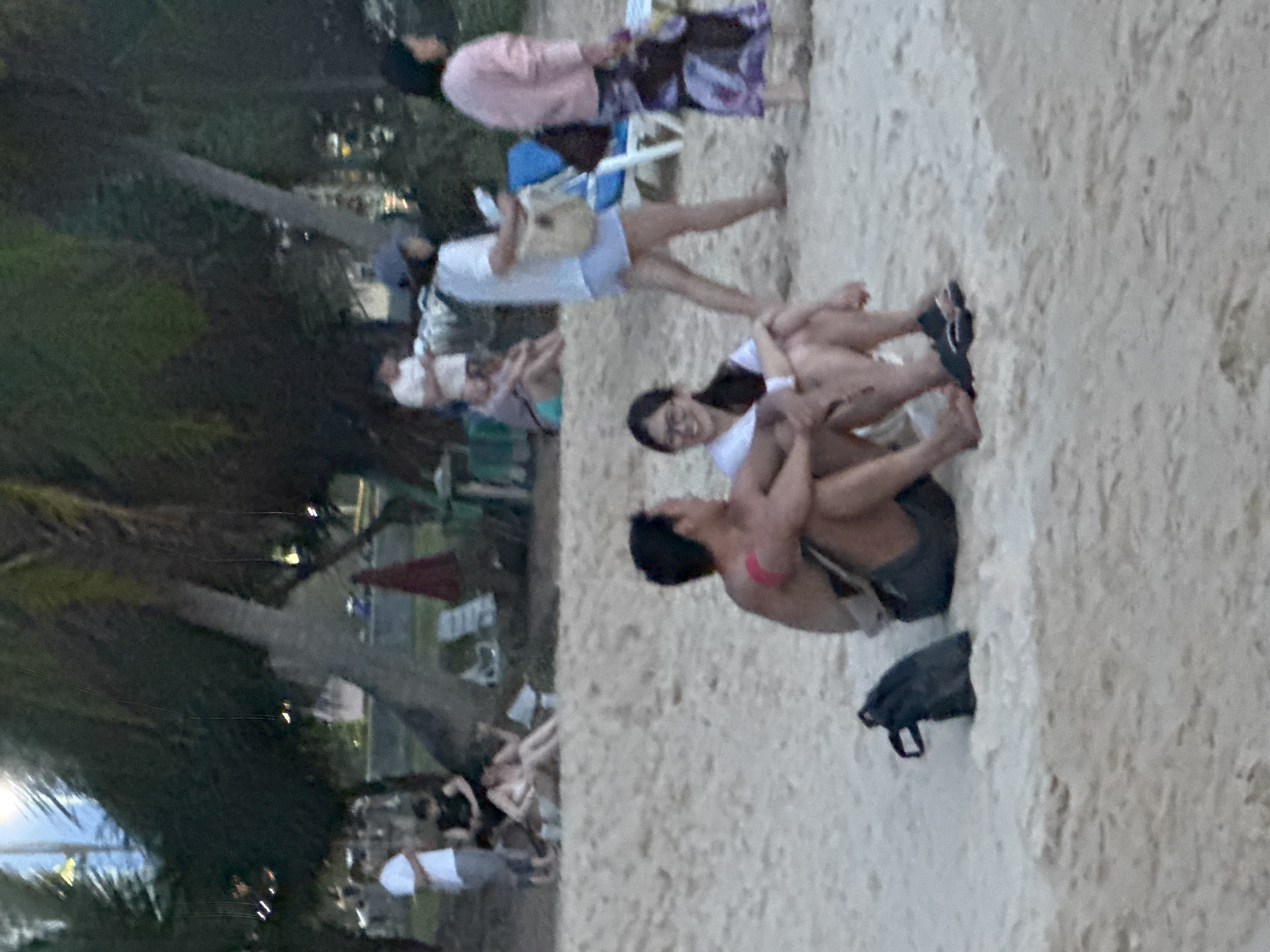 The image depicts a sandy beach scene in Karon, Thailand, likely during the late afternoon or early evening given the soft, diffused light. In the foreground, a couple is seated on the sand, facing each other and appearing to be engaged in conversation. The man is shirtless, wearing dark shorts, and has a pink band around his upper arm. The woman is wearing a white top and dark-rimmed glasses. A black bag is placed on the sand next to them.
In the mid-ground, several other people are visible. A woman in a pink top and patterned skirt is walking away from the viewer, carrying a bag. Another woman in a white top and blue shorts is walking towards the right, holding a bag. Behind them, people are seated on beach chairs and lounging on the sand, some partially obscured by palm trees. Beach umbrellas and towels are scattered on the sand.
The background shows a line of palm trees with sparse foliage, beyond which are hints of urban structures and possibly illuminated windows, suggesting proximity to a developed area. The overall atmosphere is relaxed and casual, characteristic of a beach resort setting.