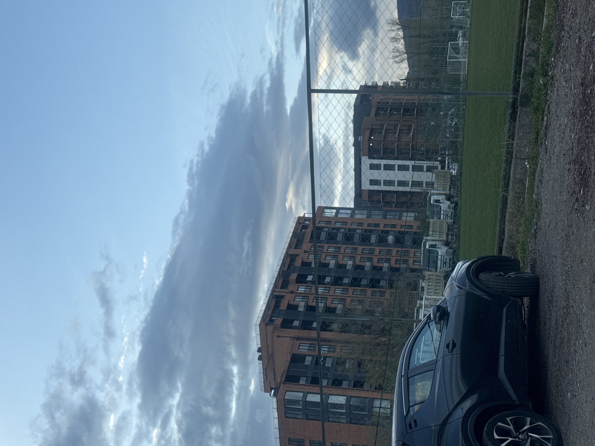 The image depicts a partly cloudy sky above the cityscape of Tbilisi, Georgia. In the foreground, a black car is parked on a gravel surface beside a chain-link fence. Beyond the fence is a grassy area, appearing to be a sports field, with what looks like soccer goals visible in the distance. Behind the field, several multi-story buildings are visible, constructed of brick and concrete. The sky shows light blue patches interspersed with grey clouds. The lighting suggests it is either early morning or late afternoon. The location is likely in a peripheral area of Tbilisi, combining residential buildings with recreational spaces. No people are visible.