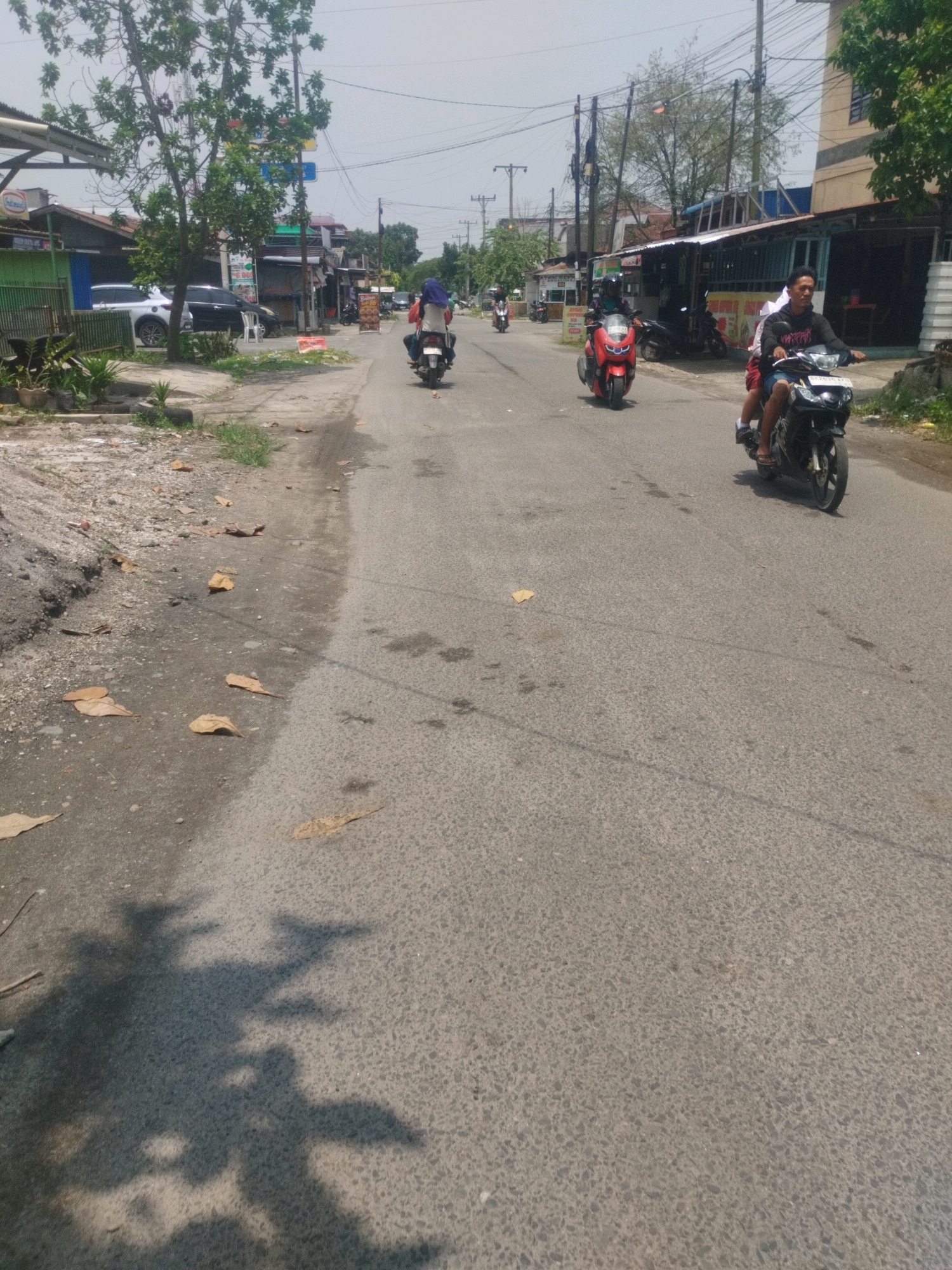 The image captures a street scene in the city of Medan, Indonesia, on a bright, overcast day. A paved road runs through a commercial area lined with buildings and shops. Several individuals are seen riding motorcycles, suggesting a mode of transportation common in the region. The foreground is dominated by the road surface, with scattered dry leaves and some visible stains or marks. The middle ground features motorcycles with riders, and the background includes buildings, trees, and utility poles, indicative of an urban environment. The overall impression is of a typical day in a bustling Indonesian city.