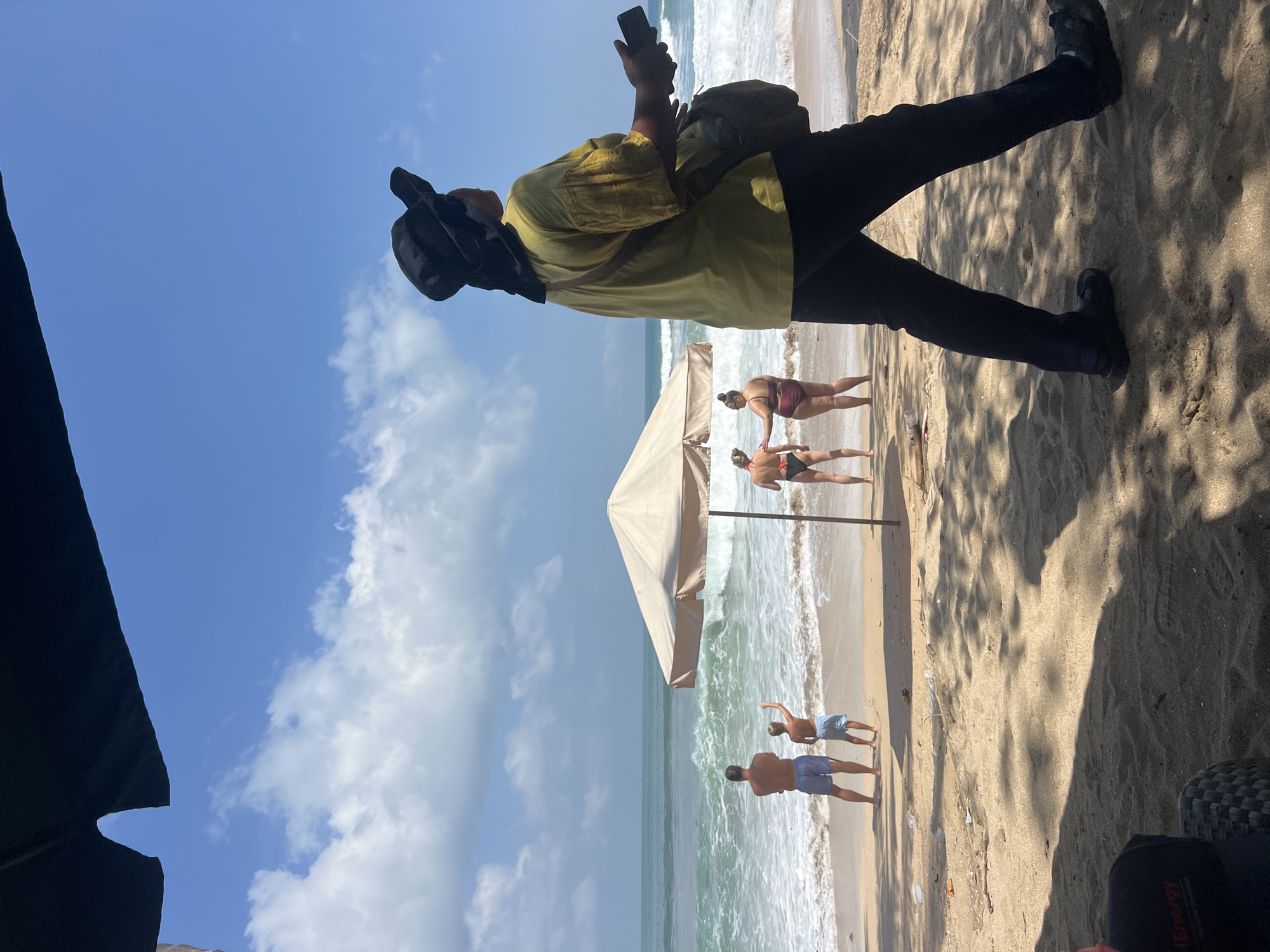 The image depicts a sandy beach in Kuta, Indonesia, under a clear blue sky with scattered clouds. In the foreground, a person wearing a black hat, a yellow shirt, and black pants is walking away from the camera, holding a mobile phone in their right hand and carrying a bag over their left shoulder. The person is casting a shadow on the sand.
Further down the beach, near the water's edge, several individuals are visible. Two adults, a man and a boy, stand facing the ocean, with the boy raising his right arm. Two women are also near the water, one with her back to the camera and the other facing away, with her left hand touching the other woman's arm. A large, light-colored umbrella stands on the sand, partially shading the area.
The ocean exhibits waves breaking on the shore, with white foam indicating active surf. The beach itself is sandy with some debris scattered on its surface. The overall scene suggests a relaxed day at the beach with recreational activities taking place.