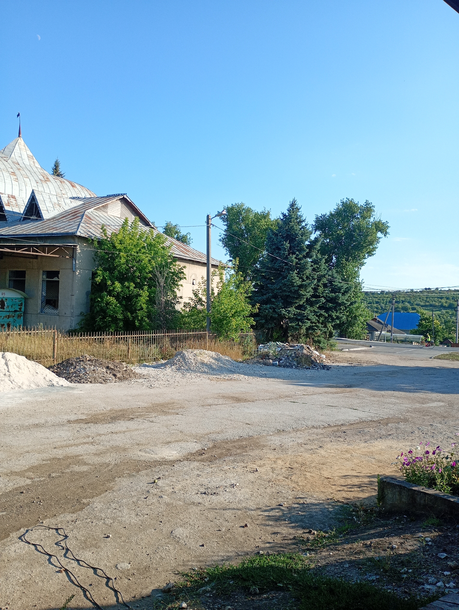 The image shows a sunny day in Sîngerei, Moldova, with a clear blue sky and a crescent moon visible. The foreground is a dusty dirt road with piles of construction materials, including sand and gravel. To the left, an old, weathered building with a corrugated metal roof and a covered entrance is visible, with a rusty metal fence in front. Trees and bushes surround the building. In the middle ground, several large evergreen trees stand prominently. Further back, a road winds uphill, with houses and trees on either side. A person on a bicycle is visible on the road, heading uphill. The background consists of rolling hills covered in vegetation.