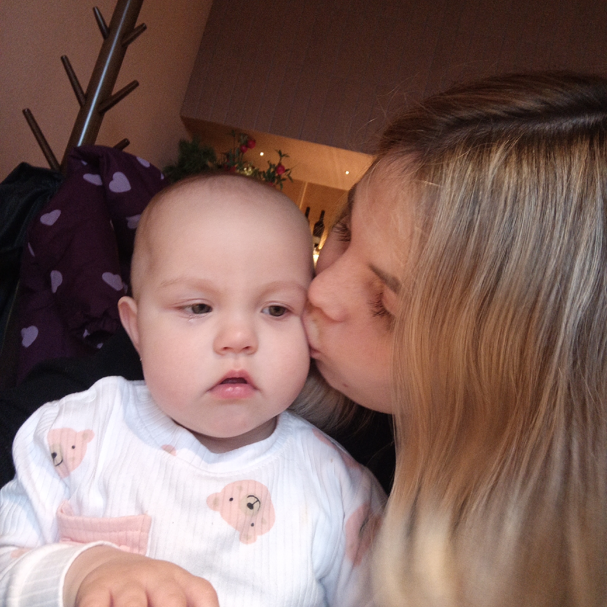 An indoor, close-up shot captures an adult kissing a baby on the cheek. The adult, positioned on the right, has blonde hair and is leaning in, partially obscuring their face with the baby's head. The baby, in the foreground, faces slightly away from the adult and towards the viewer, displaying light skin and dark eyes. A small tear is visible in the corner of the baby's left eye, giving their expression a melancholic or solemn quality. The baby wears a white, ribbed long-sleeve top adorned with multiple pink teddy bear designs.
The background reveals elements of an indoor setting, possibly a home. To the left, a wooden coat rack with several pegs is visible, from which hangs a dark purple garment patterned with light purple hearts. Further into the background, some decorative green foliage interspersed with red accents, possibly artificial berries or flowers, can be seen, along with what appear to be bottles on a shelf. The scene is lit with warm, artificial light, suggesting it could be any time of day or evening indoors. There is no visible text.