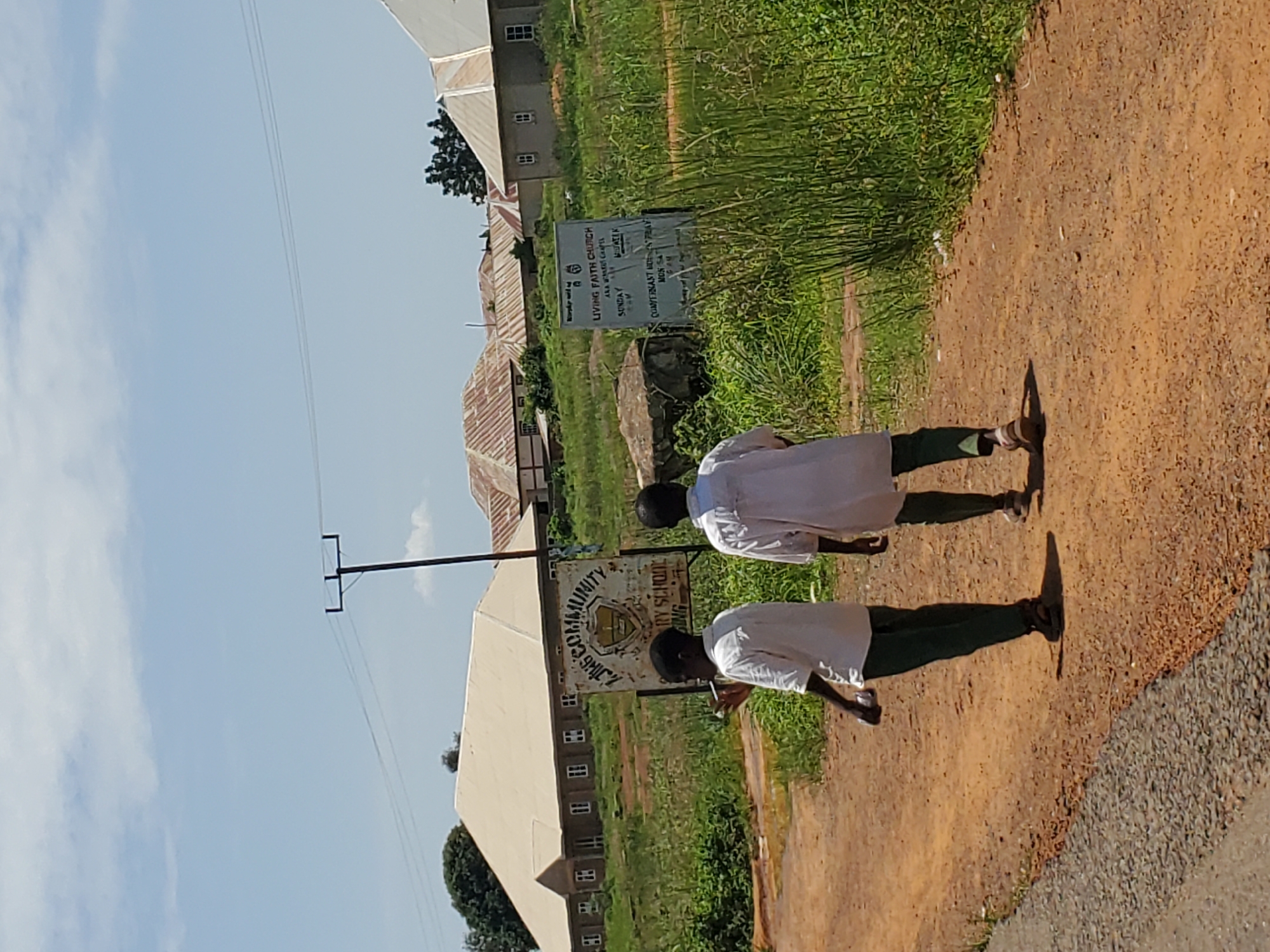 This media file depicts a scene in Mangu, Nigeria, under a clear blue sky with scattered clouds, suggesting it is daytime. Two individuals, likely students given their white uniforms, are walking away from the camera on a dirt path. The path is bordered by a grassy area on the left and a darker, gravelly surface on the right. In the background, several buildings with corrugated iron roofs are visible, indicative of a rural or semi-urban setting.
A signpost stands prominently in the middle ground, displaying text that includes "LIVING FAITH CHURCH," "AKAWATCHA," "MATEK," and "COMMUNITY RY SCHOOL." It also lists service times for "SUNDAY" and "MON SAT." Another signpost is partially visible to the left, with "COMMUNITY" clearly legible. The presence of these signs suggests a community area, possibly near a church and a school. The overall impression is one of a typical day in a Nigerian community, with children on their way somewhere and religious and educational institutions present. The shadows cast by the individuals indicate the sun is relatively high in the sky.