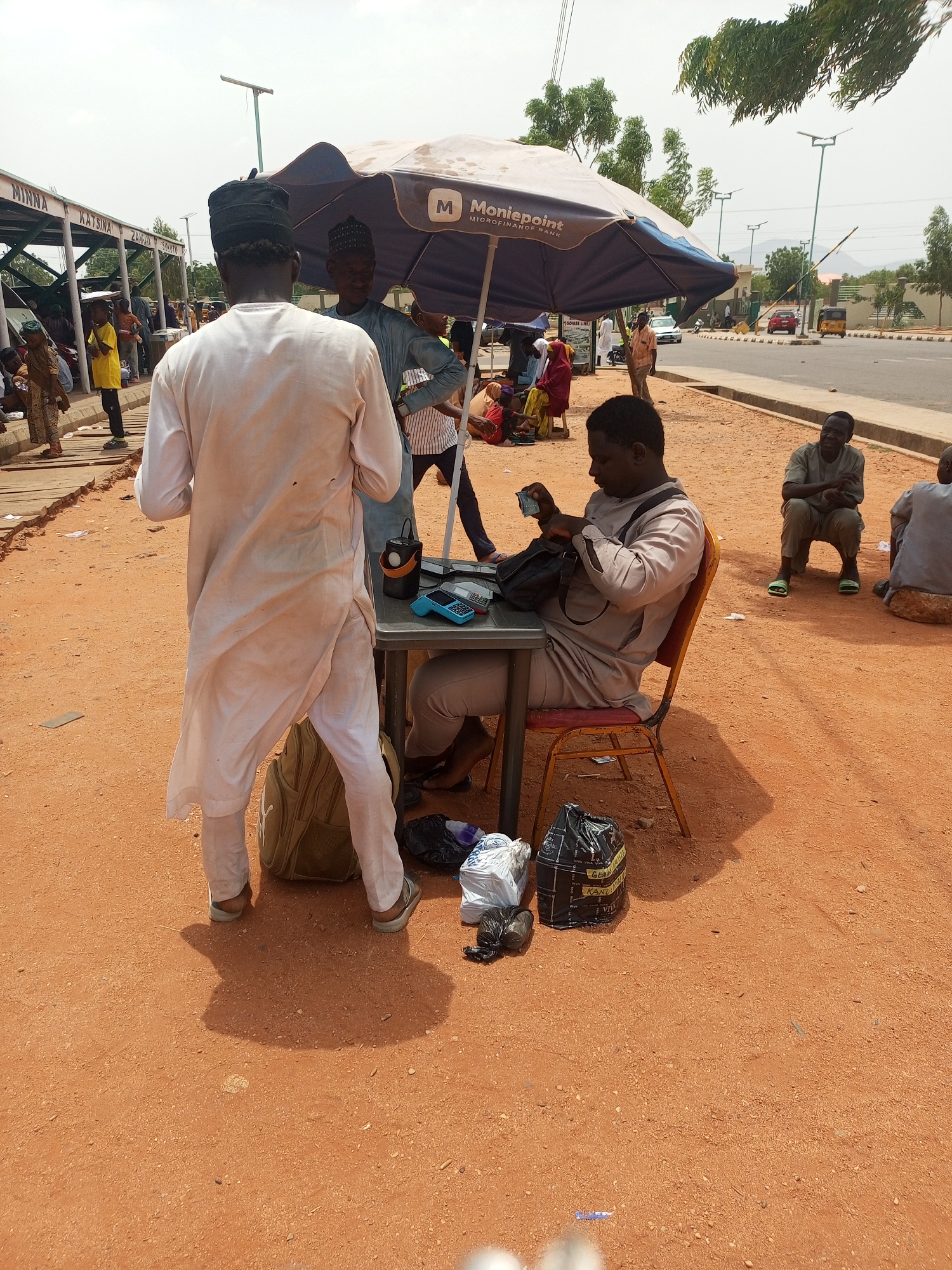 The image depicts an outdoor scene in what appears to be a city, possibly in Africa, given the people's attire and the Moniepoint umbrella. A man is seated at a small table, counting money from a black bag, under the shade of the Moniepoint umbrella. The individual wears a light-colored, casual suit. There is another man standing in front of the table with his back to the camera, wearing a long, white robe and a black cap, with a backpack at his feet. A third man stands by the table. The ground is reddish-brown, likely dirt or sand. There are several bags near the table. In the background, there are other individuals, buildings, and a street with vehicles, with "MINNA" appearing on a structure in the far background. One man sits on the ground on the right, and another is beside him. The environment appears to be a somewhat busy public area.
