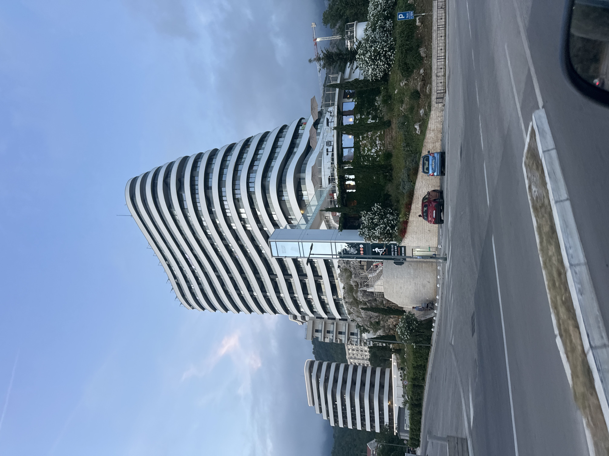 This image depicts a modern architectural building in a hilly, possibly coastal, setting during daytime. The central subject is a multi-story building on the left, characterized by its distinctive wavy, horizontal balconies that alternate between light white/grey and dark glass. It appears to be a hotel or apartment complex.
To the right, a two-lane asphalt road with white markings runs through the foreground. Two cars are parked near the road: a red hatchback and a blue hatchback. The surrounding landscape features abundant greenery, including bushes with white flowers and trees, climbing up the hillsides. In the background, green, forested hills are visible, and a construction crane stands out on a distant slope. Another smaller building with similar wavy architecture can be seen behind and to the right of the main structure.
The sky is a bright, clear blue with a few scattered, soft clouds, suggesting a pleasant daytime, possibly morning or late afternoon, given the light quality.
Visible text includes a blue sign with a white 'P' symbol for parking and the word "TAXI". Near the main building's entrance, a larger sign is partially visible, with a clear smaller sign beneath it that reads "WHERE TO GO FOOD & BAR LA ROCHE". The scene is static, with no discernible human activity. The overall impression is one of a modern resort or residential area nestled into a natural, hilly landscape in Boreti, Montenegro.