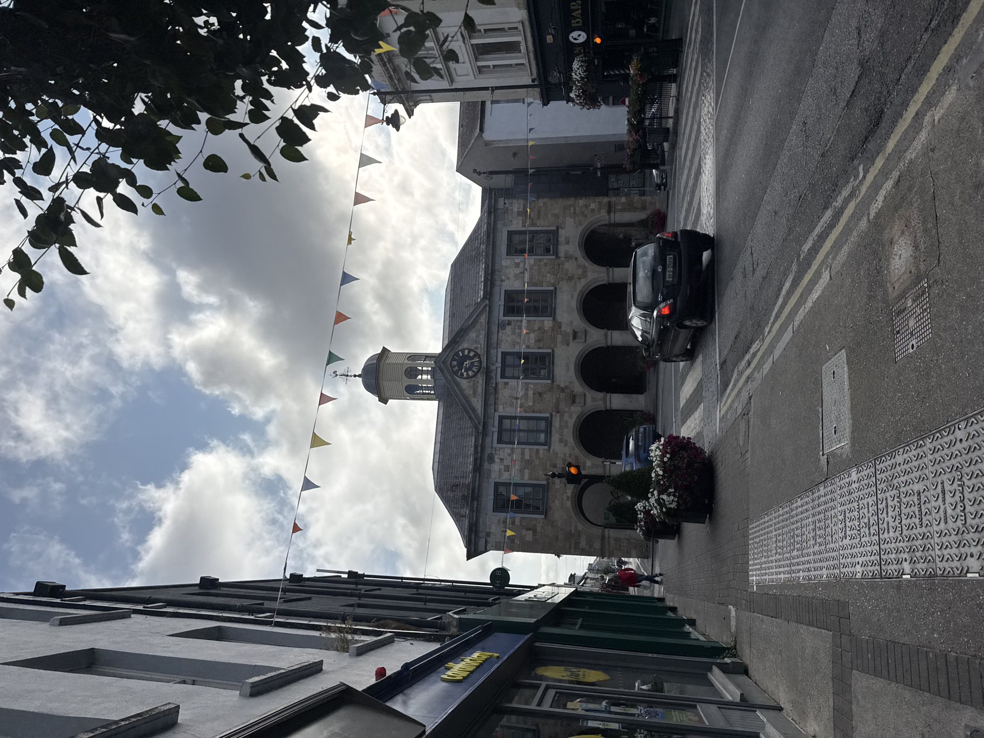 The image shows a street scene in Clonmel, Ireland. The focal point is a historic stone building with arched entrances, likely a town hall or market house, which features a clock tower. Colorful triangular flags are strung across the street above the building. In the foreground, a black Kia car is driving on the right side of the road, passing by a pedestrian sidewalk. The sidewalk is lined with buildings, one of which has a green facade with a sign that reads "Card Factory." There are also planters with flowers on the sidewalk. The sky is partly cloudy, with sunlight filtering through. The street has double yellow lines indicating parking restrictions and a pedestrian crossing is visible in the distance. A person in a red jacket is walking on the sidewalk.