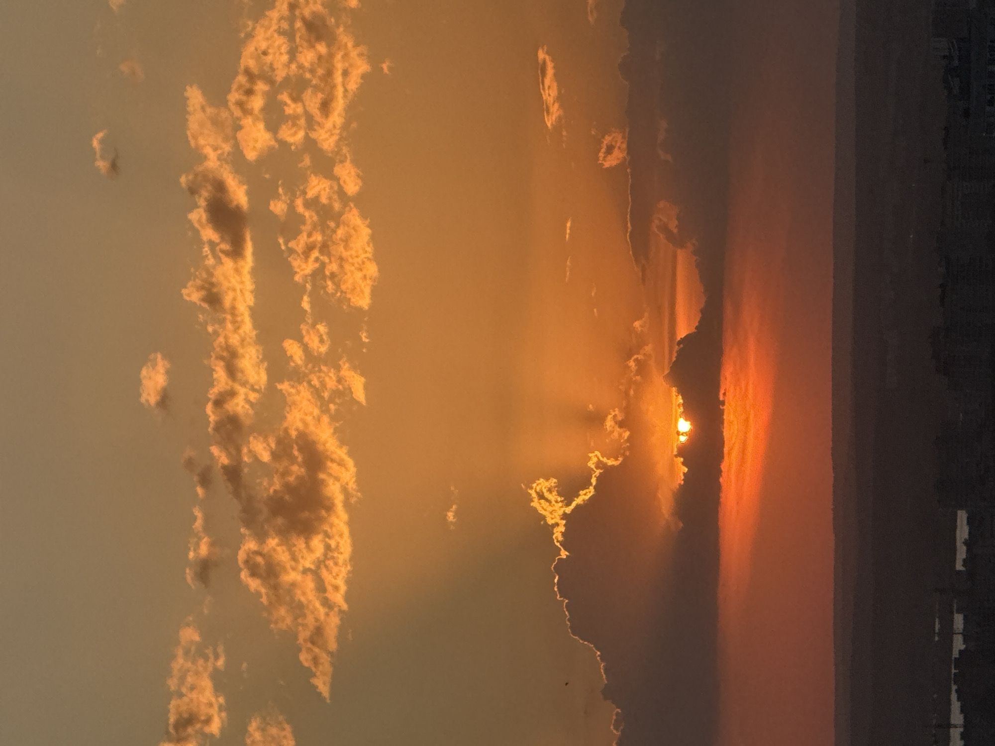 A vertical, eye-level, outdoor shot shows a sunset over the city of Perm, Russia. The sky is filled with clouds of varying sizes, some illuminated with orange and yellow light from the setting sun. The sun itself is partially obscured by a darker cloud bank near the horizon, but its rays stream out in several directions. The horizon is somewhat obscured by haze. A dark mass, potentially a cityscape or treeline, is visible at the bottom right of the image.