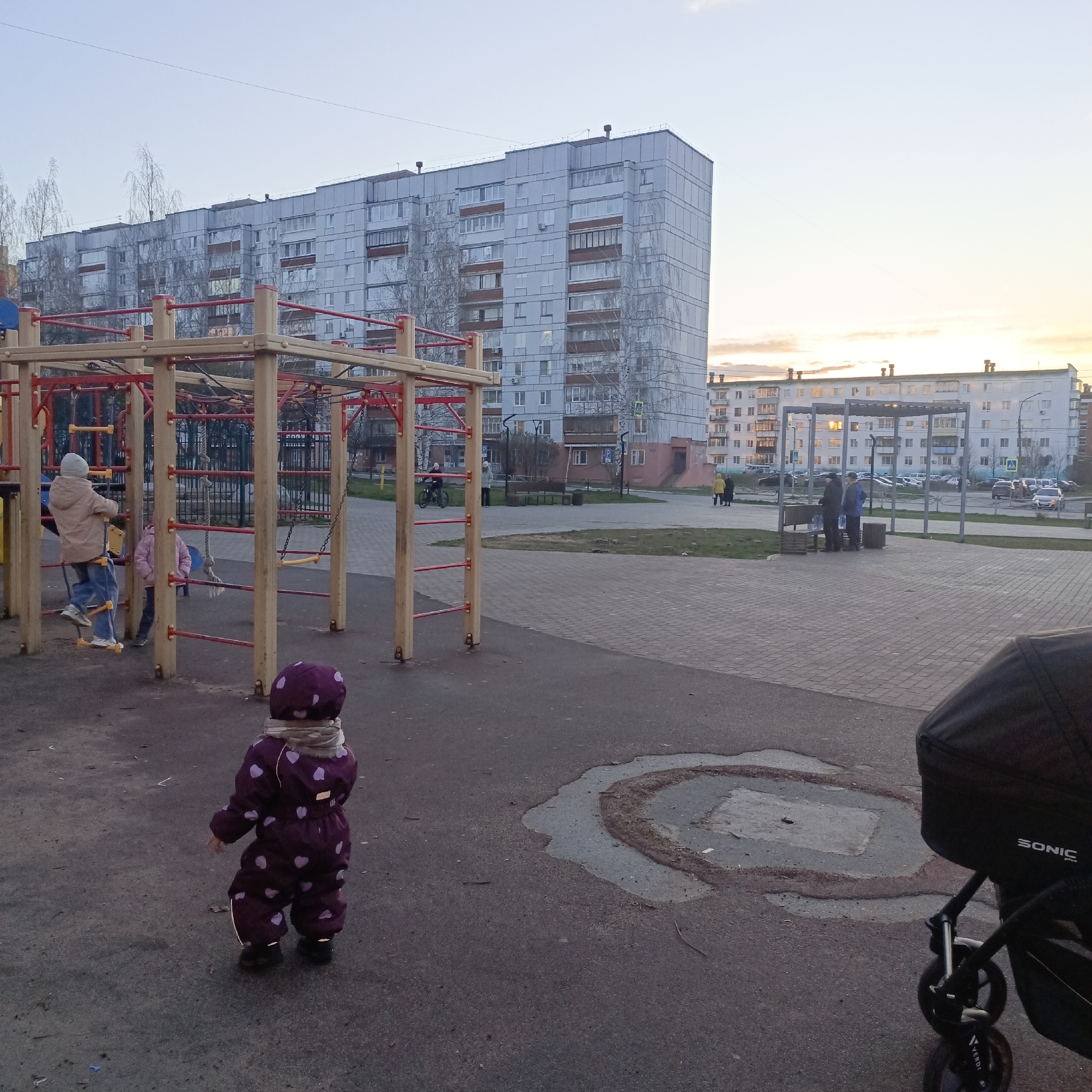 A wide shot captures a residential area playground at dusk, characterized by the soft, warm glow of the setting sun on the horizon. In the immediate foreground, a toddler, dressed in a purple winter suit with light heart patterns and a patterned scarf, stands with their back to the camera on an asphalt surface. To the right, part of a black baby stroller is visible, with the brand names "SONIC plus" and "VERDI" faintly legible on its fabric and wheel, respectively. The asphalt shows signs of wear, with cracks and a circular discolored patch.
The midground features a large wooden and red-metal playground structure. On the structure, two children are present: one in a light-colored jacket and blue jeans is actively climbing a ladder-like section, while another child in a pink jacket is partially obscured. Beyond the playground, a paved area with patches of grass leads towards apartment buildings. Several adults are in the mid-distance; two are conversing near a bench under a metallic pergola, and others are walking or riding a bicycle further back.
In the background, two prominent apartment buildings dominate the skyline. The left building is a tall, light-grey structure with numerous windows and balconies. To the right, a shorter, white building reflects the soft, golden light of the setting sun on its windows. Bare-branched trees are scattered between the playground and the buildings. The sky above is a gradient of light blue, transitioning to soft orange and pink hues near the horizon, indicating the evening hour. The overall scene depicts a casual, everyday moment in a suburban or urban residential complex.