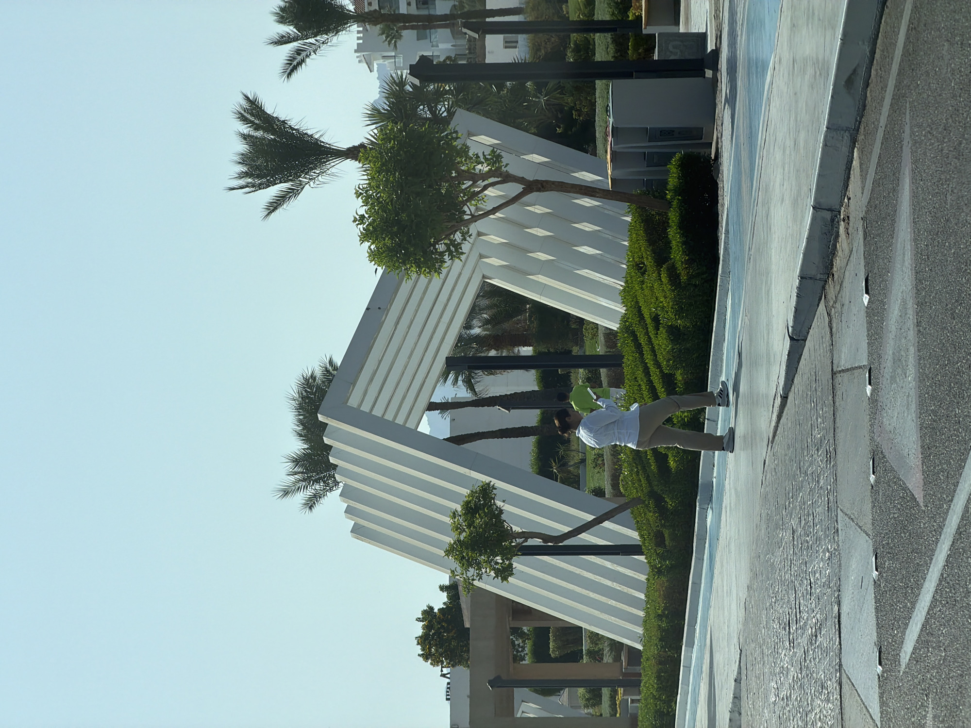 A woman in a white shirt and tan pants walks on a paved path in a resort area, holding a green balloon. Behind her is a modern, abstract white structure with angled slats, surrounded by lush green bushes and trees. Palm trees and white buildings are visible in the background, suggesting a warm, sunny climate. The overall scene depicts an outdoor public space in Sharm El Sheikh, Egypt, likely a resort or hotel grounds, designed for leisure and relaxation.