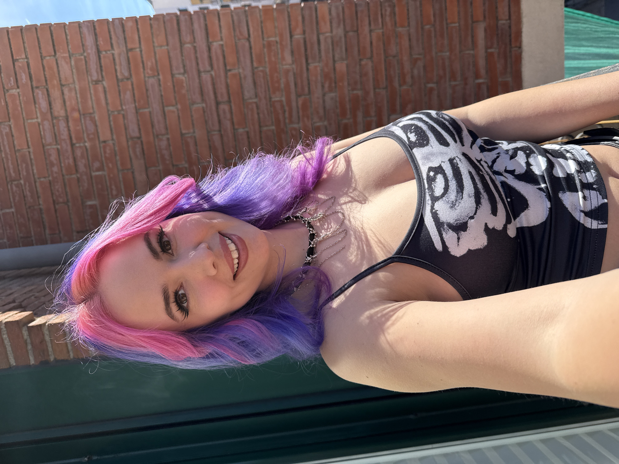 The image is a selfie of a woman with pink and purple hair, wearing a black and white top with a skeleton-like print. She's smiling at the camera. She is wearing a black chain necklace. Behind her is a brick wall. The location is Madrid, Spain.