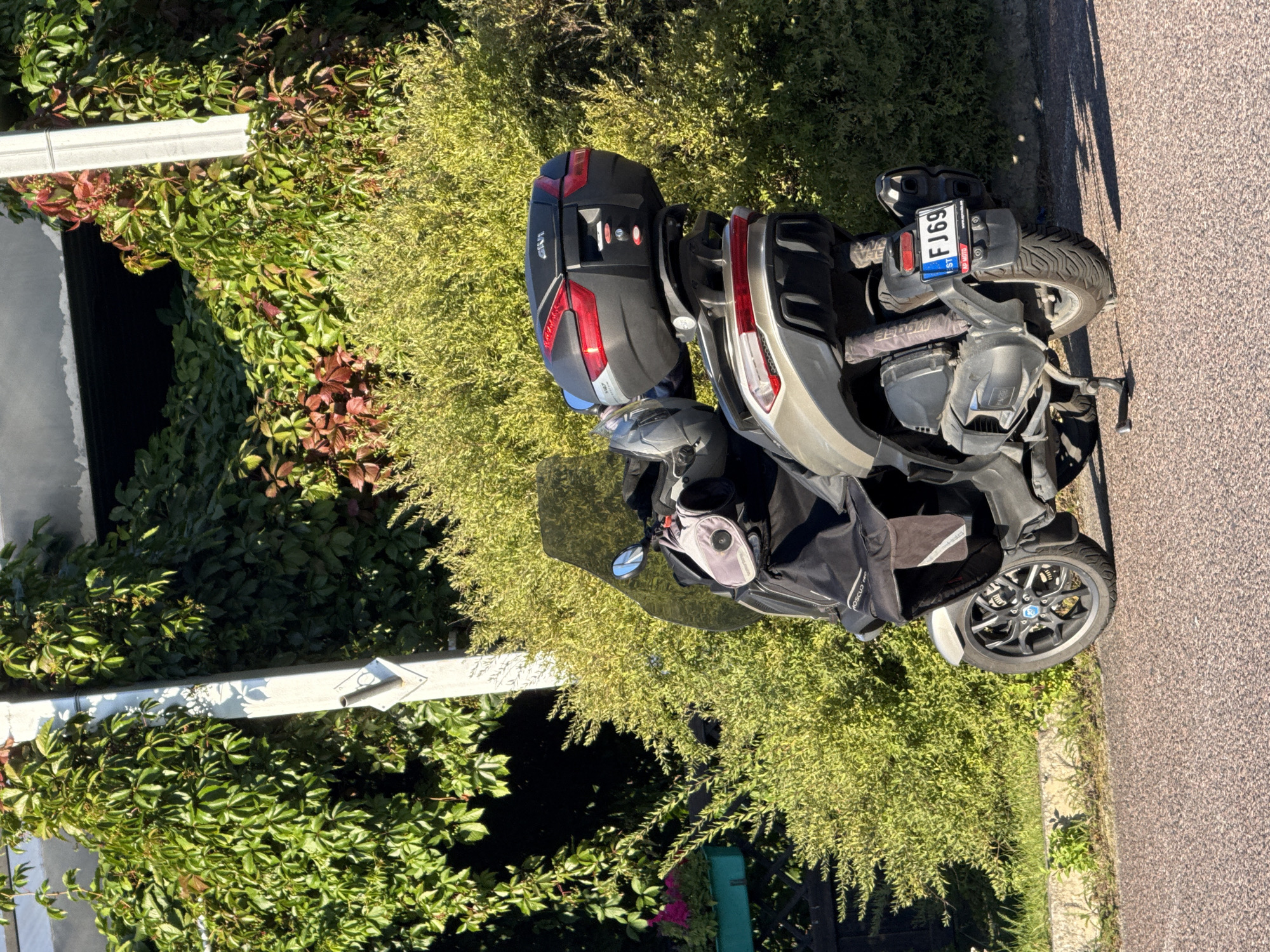 A three-wheeled scooter is parked on the side of a paved road in front of a lush green bush and a building with ivy-covered walls. The scooter is facing away from the camera and has a large black storage box on the back. A license plate that reads "FJ69" is visible on the rear fender. The scene appears to be in Tallinn, Estonia, given the style of the license plate and the presence of a building that is typical of Estonian architecture. The scooter is covered with a black fabric, possibly a rain cover or a bag. The scooter's mirror is reflecting a blue sky and some green foliage. The lighting suggests it is a sunny day.