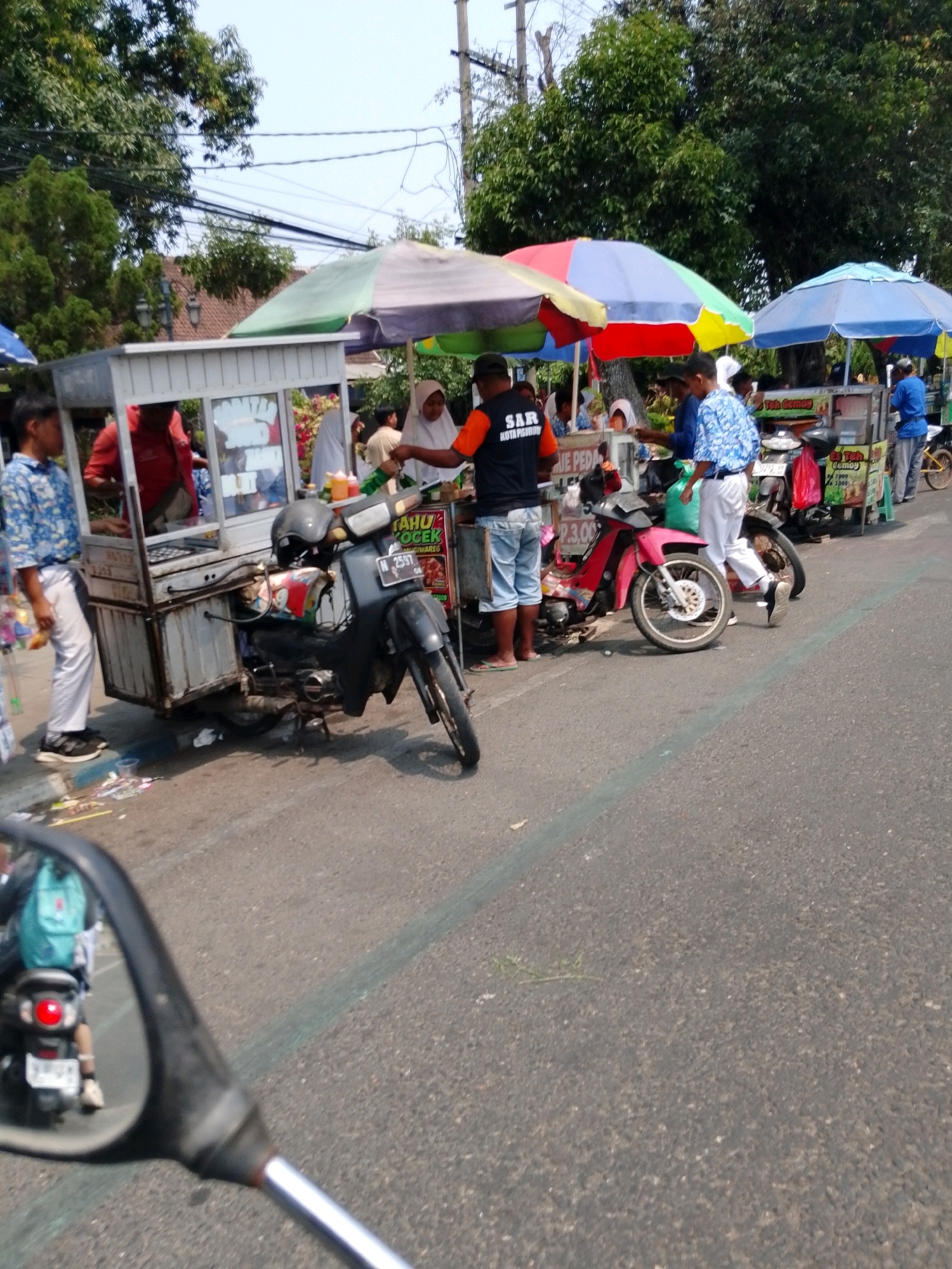 The image depicts a street scene in Pasuruan, Indonesia, likely during daylight hours. A food cart, possibly selling "Tahu Goreng" (fried tofu) and "Teh Gemoy" (a type of tea), is parked alongside a road. The cart is covered by a multi-colored umbrella. Several people are gathered around the cart, with some appearing to be customers and others vendors.
In the foreground, a black motorcycle is parked next to the food cart. A man wearing a blue and white patterned shirt and white pants is standing near this motorcycle, holding an item. To the left of the food cart, a young man in a blue and white patterned shirt and white pants stands with his hands in his pockets. Behind him, a red-shirted figure is partially visible within the cart.
Further down the road, another food cart is visible, also with an umbrella. A man in a blue shirt is seen next to this cart, and another person in a blue outfit stands near a yellow bicycle. A red motorcycle is parked in front of the second food cart.
The environment includes a paved road with a faint green line marking. Trees with lush green foliage line the background, and power lines are visible overhead. Debris is scattered on the side of the road near the first food cart. The image appears to be taken from the perspective of someone on a motorcycle, as a rearview mirror with a reflection of a motorcycle is visible in the bottom left corner.