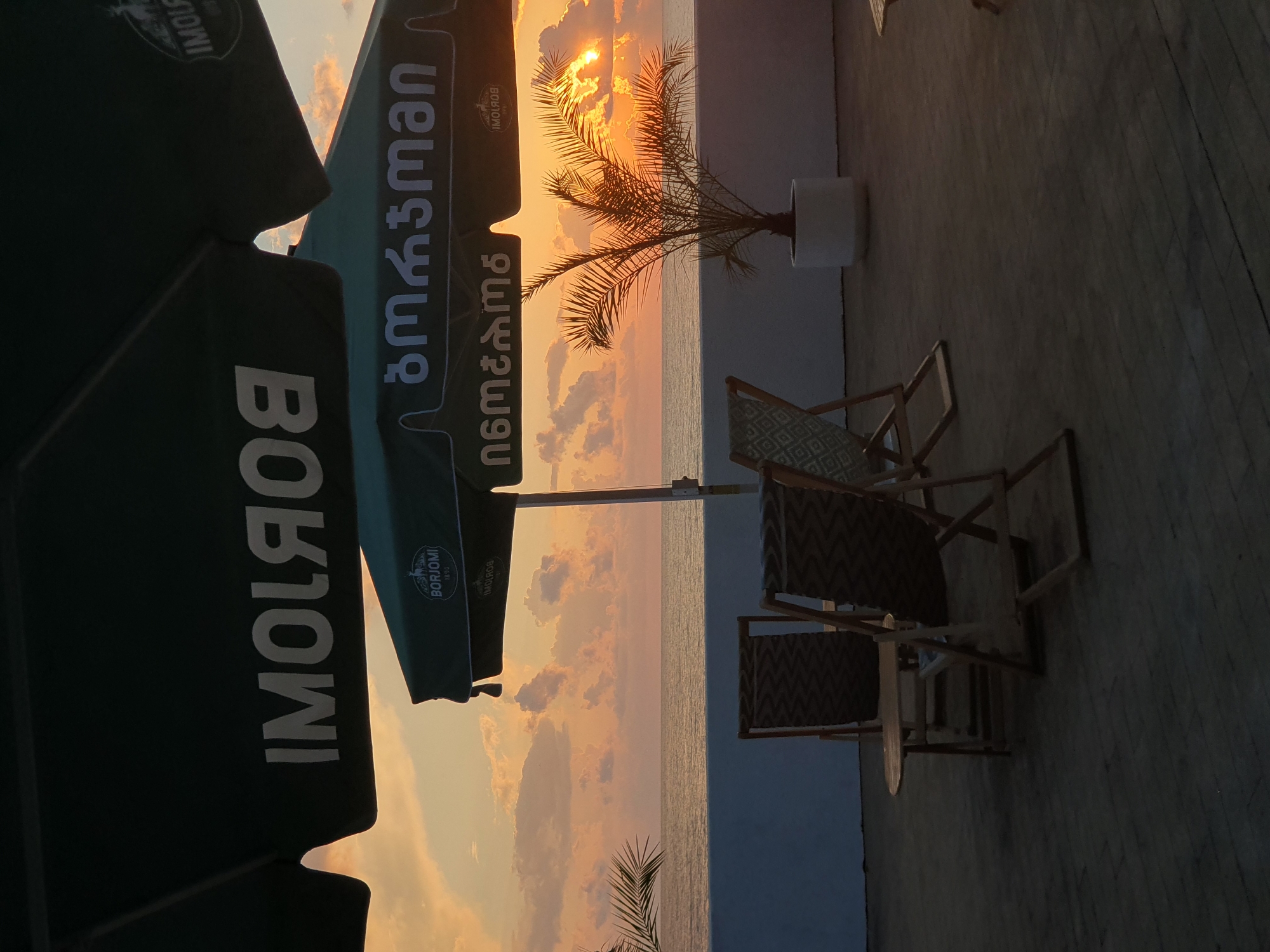 The image depicts a sunset scene at a waterfront cafe, likely in Batumi, Georgia. Two green umbrellas with white lettering are visible, one partially obscuring the sky and featuring the text "IMOLЯOB". The other umbrella, positioned lower, displays the logo of "BORJOMI" along with Georgian script that appears to read "ბორჯომი" and "ინეტირება". In the foreground, two wooden deck chairs with patterned cushions and a small wooden table are set up on a wooden deck. To the right, a potted palm tree stands in a white planter against a white wall. Beyond the wall is the calm sea, reflecting the warm hues of the sunset sky, which is filled with orange and pink clouds. The overall ambiance suggests a relaxed outdoor seating area overlooking the Black Sea at dusk.