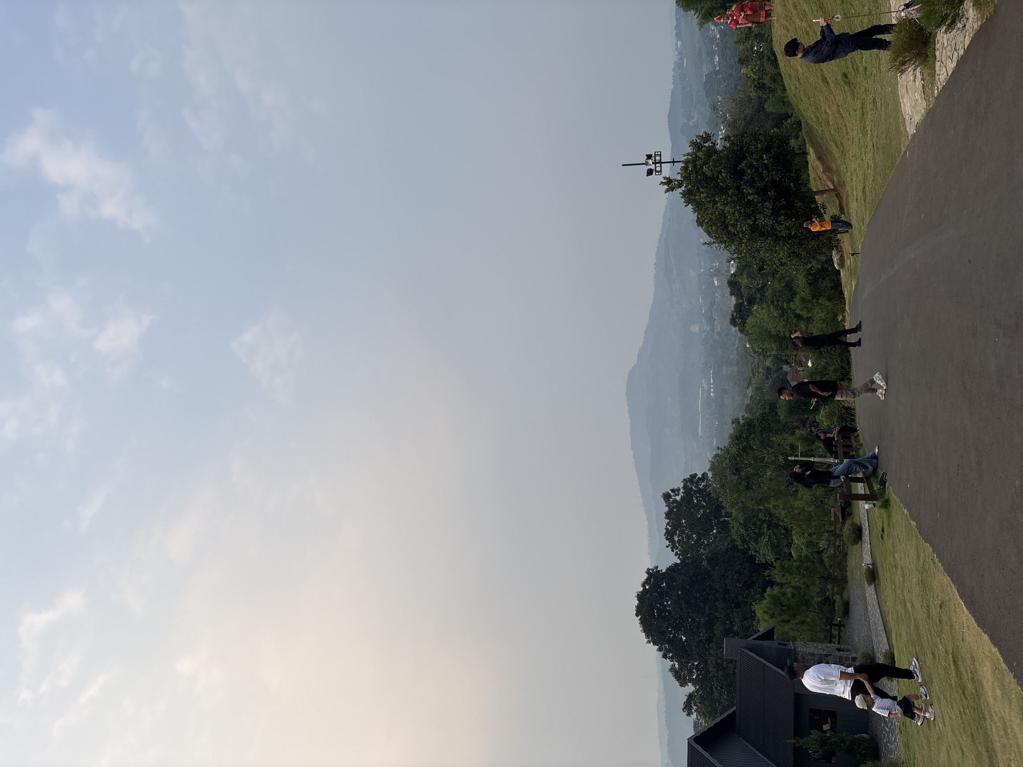 The image depicts a scenic outdoor setting in Babakan Madang, Indonesia, likely during the day given the bright sky with scattered clouds. The scene features a group of people engaged in various activities. In the foreground, two individuals are interacting near a dark-colored modern building with a pitched roof. Further up a grassy slope, several other individuals are scattered along a paved pathway that winds through the landscape. One person is standing on the grass, holding what appears to be a pole. Another is seated on a bench, and a few others are walking or standing. In the background, lush green trees cover the rolling hills, with a hazy view of distant mountains and scattered buildings suggesting a rural or semi-rural environment. A light pole with multiple fixtures stands on the slope. The overall atmosphere appears relaxed and casual, with people enjoying the outdoor environment and perhaps a gathering or outing. There is no visible text in the image.