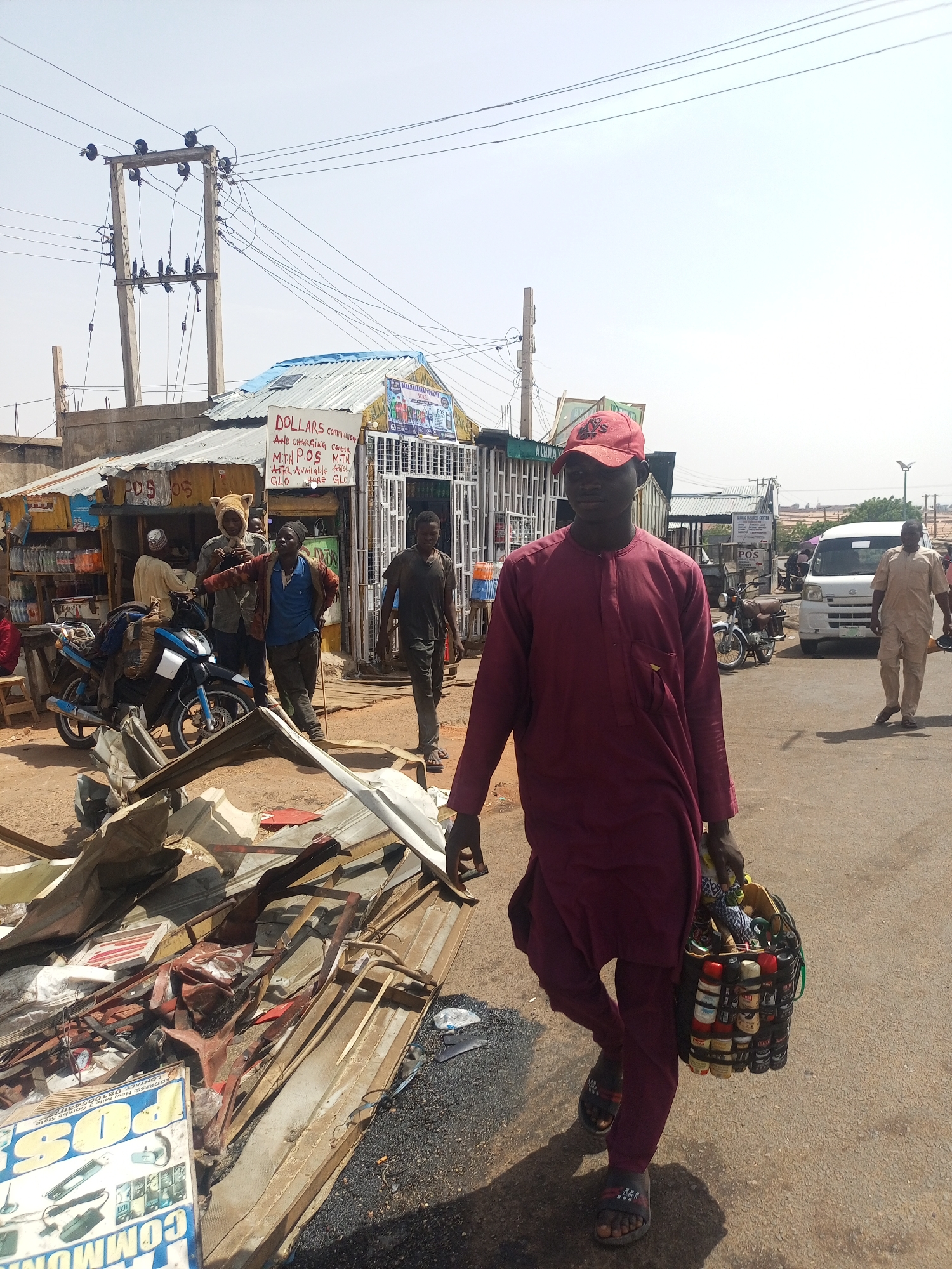 The image captures a street scene in Gombe, Nigeria. A man in a maroon outfit and red cap walks on the right side of the frame, carrying a bag filled with cylindrical containers. In the background, a small shop stands under an overcast sky, with signs reading "DOLLARS Communication and Charging Center, MTN POS, MTN Airtime Available Here, GLO". Other people are present near the shop, and motorcycles are parked nearby. Debris is scattered on the ground in the foreground, adding to the scene's complexity. Utility poles with wires extend overhead, contributing to the urban environment. Vehicles are visible further down the road, suggesting an active street.