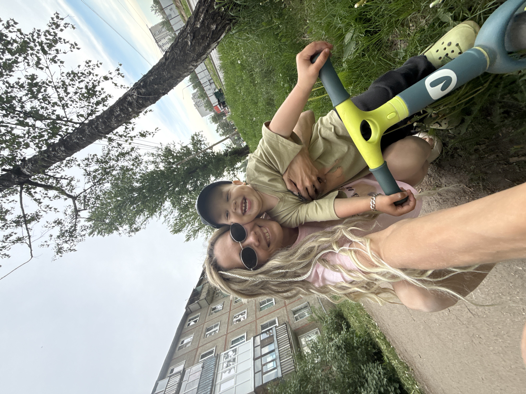 A mother and child are taking a selfie in Rusanovo, Russia. The photo is taken at an angle, facing upwards towards the sky, partially capturing a tall birch tree and multi-story apartment buildings. The mother, wearing round sunglasses and blonde hair in braids, is holding the phone. The child is smiling and hugging the mother. They are sitting in the grass, alongside a child’s toy scooter.