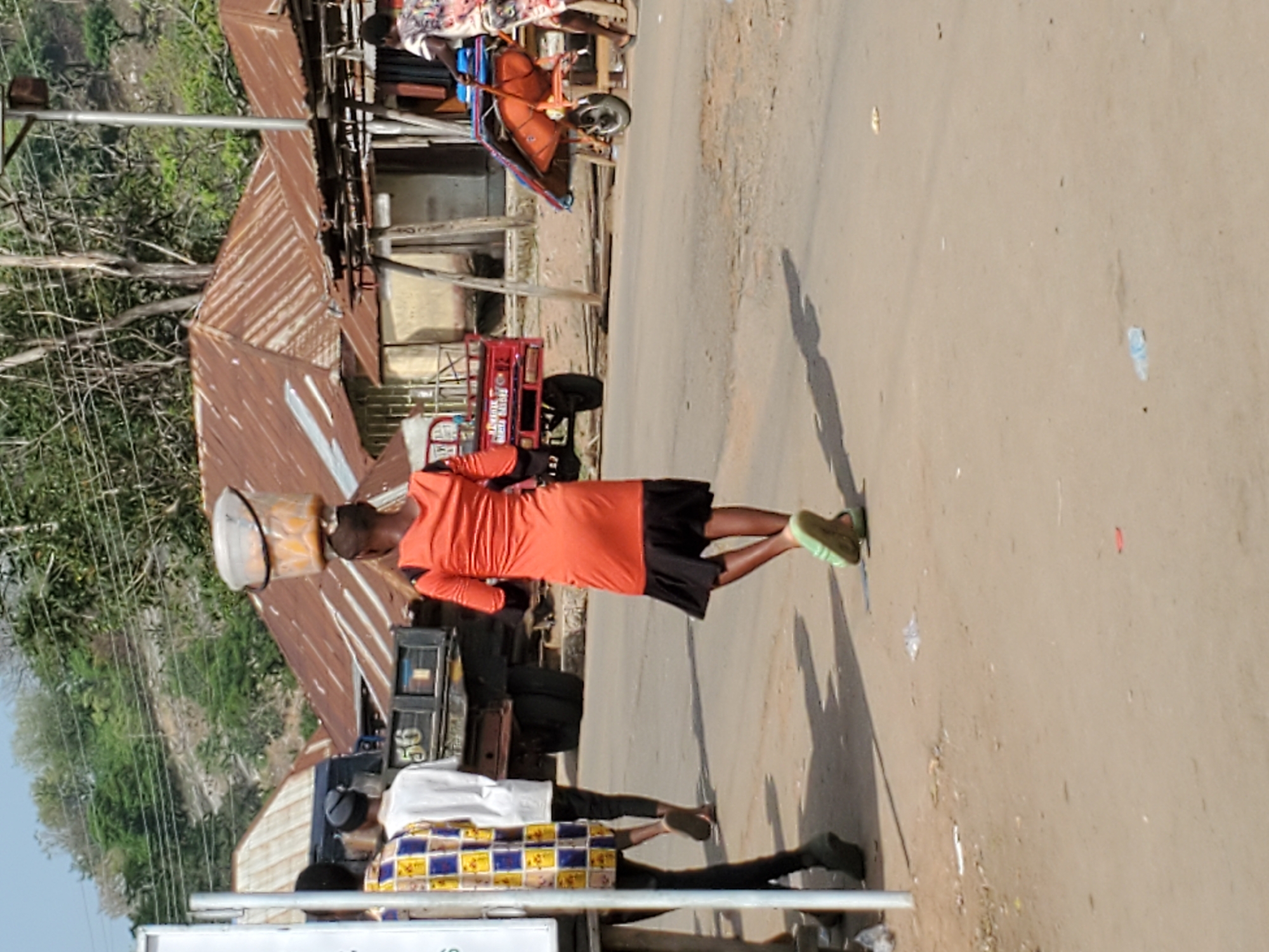 This image captures a bright, sunny daytime scene on a street in Mararaba, Nigeria. The primary focus is a person, likely a young woman, dressed in a bright orange top and black skirt, walking along a dusty or unpaved road. She is carrying a large, light-colored pot or basin balanced skillfully on her head, a common method of carrying goods in the region. Her shadow is cast long on the ground behind her, indicating the sun is relatively low, possibly late morning or afternoon. She wears light green shoes.
In the foreground, another person is partially visible, wearing a white shirt and a distinct yellow, blue, and white patterned wrapper or skirt, standing near a structure. Behind them, a red tricycle, commonly known as a "keke napep" in Nigeria, with a cargo bed is parked or driving. Text visible on the side of the tricycle includes "SUPER HERO" and possibly "TRICYCLE" below it.
The background features several buildings with rusty corrugated iron roofs, characteristic of local architecture. Lush green trees and vegetation are visible behind these structures, suggesting a somewhat rural or semi-urban environment. Utility poles and overhead power lines are also discernible. The street surface appears to be packed dirt or gravel with some debris. The overall atmosphere suggests daily life and commerce in a bustling community.