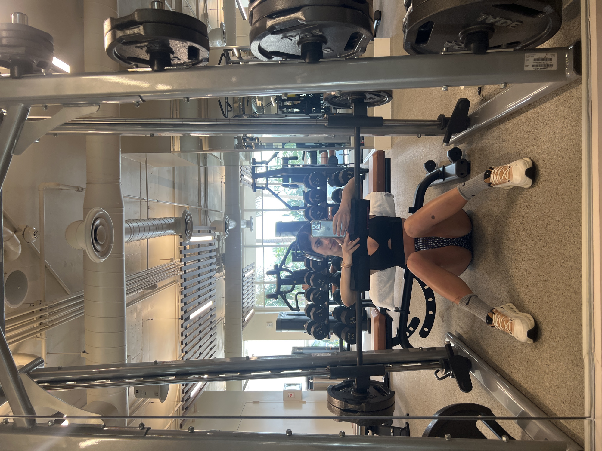 A woman is taking a selfie while lying on a bench press in a gym in Miami Beach, United States. The gym has multiple weight machines, dumbbells, and a mirrored wall. The ceiling has exposed pipes and ventilation systems. The woman is wearing a black top, patterned shorts, and gray socks with white and tan sneakers. She is holding a smartphone in her right hand and looking at the screen.