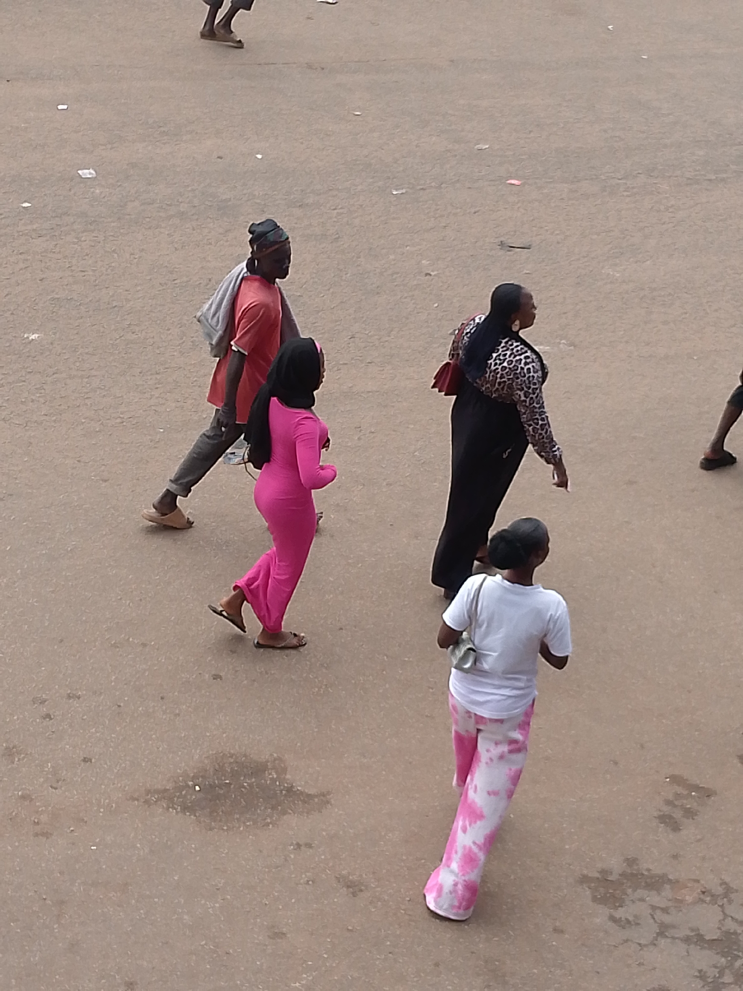This image depicts a street scene in Bukuru, Nigeria, from an elevated perspective. A man wearing a red t-shirt and dark pants is walking with a gray bag draped over his shoulder. To his right, a woman in a bright pink full-length dress and a black hijab is also walking. Further to the right, another woman is seen wearing a leopard-print top, black pants, and carrying a red purse. In the foreground, a younger woman in a white t-shirt and tie-dye pink and white pants is walking away from the camera, carrying a light-colored bag. The ground appears to be a dusty, paved surface with scattered debris.