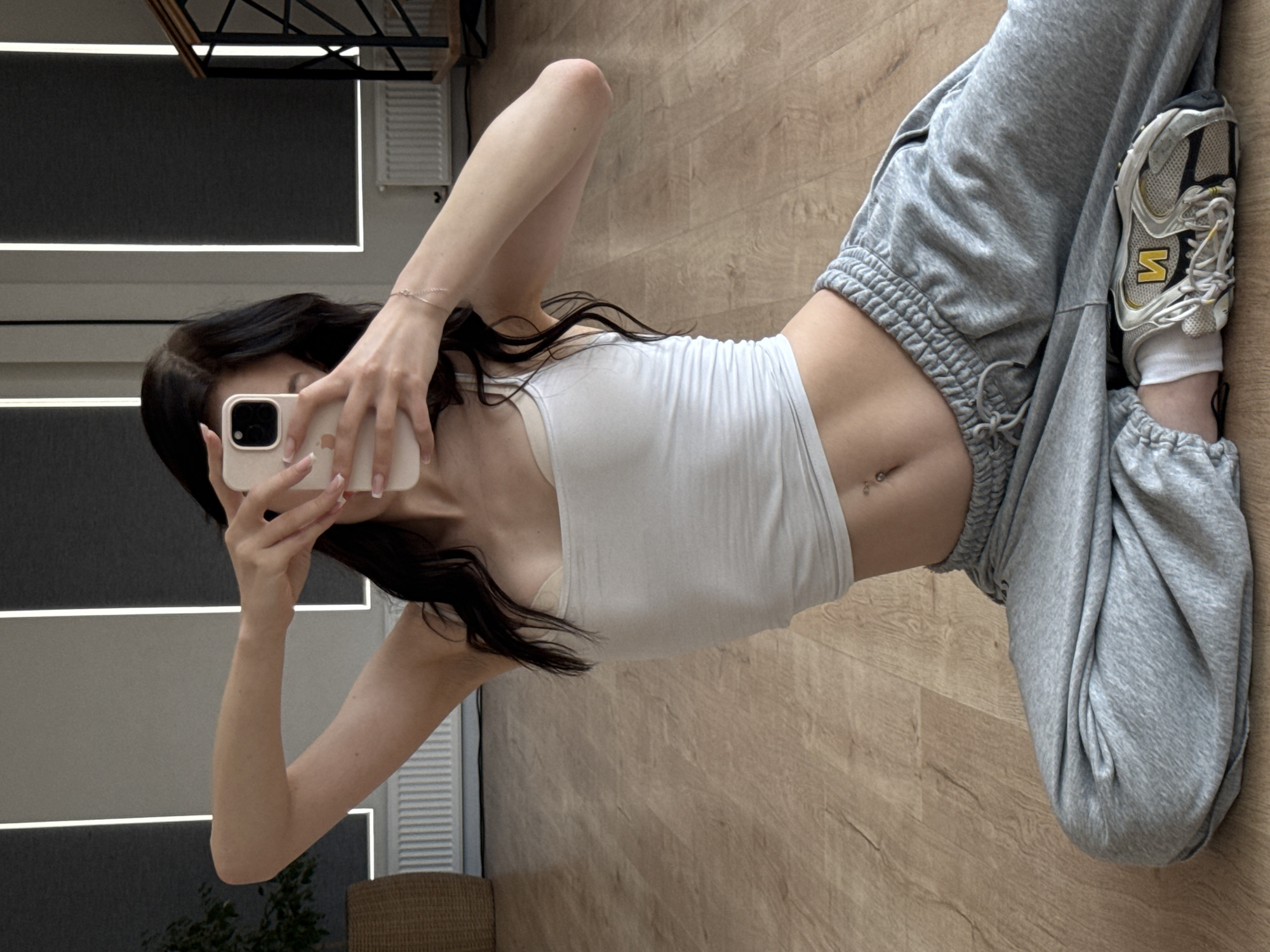 This media file shows a person taking a mirror selfie in a room with light wood flooring. The person is lying down, with their legs bent and one foot resting on the other. They are wearing a white tank top, grey sweatpants, and athletic shoes. The tank top exposes their midriff, revealing a belly button piercing. The room appears to be a home, with a white vent visible on the wall in the background, and a glimpse of furniture. The lighting suggests it is daytime. The image is an indoor shot, and there is no visible text.