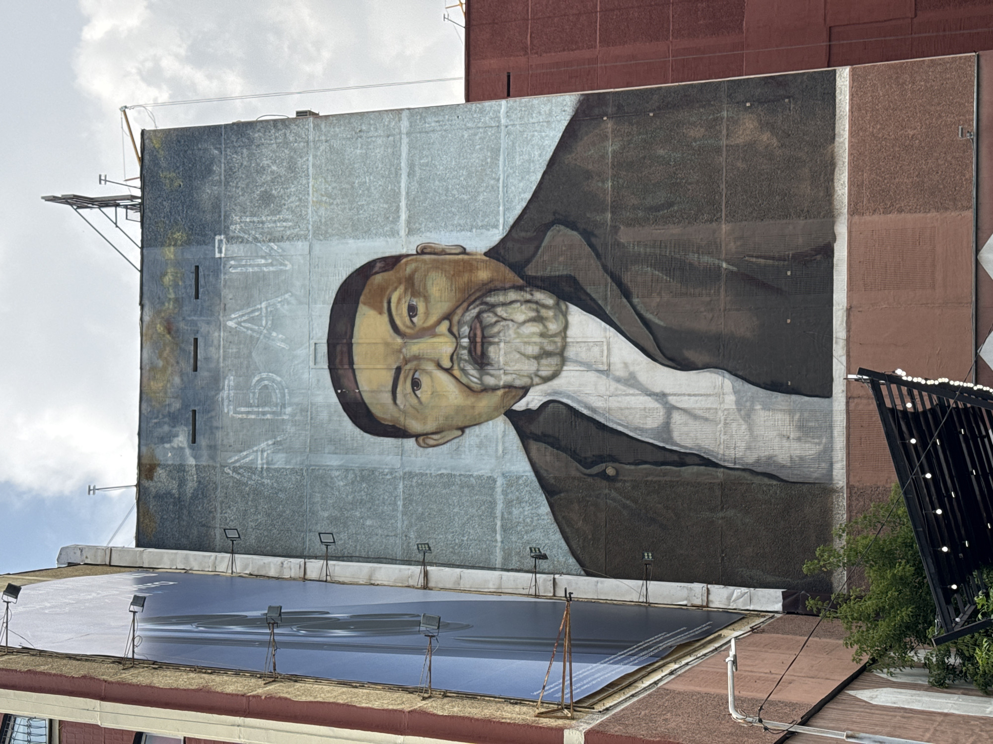 A large mural of a man with a beard is painted on the side of a building in Almaty, Kazakhstan. The man is wearing a dark jacket over a white shirt and has a stern expression. The mural is located in an urban environment, with other buildings visible in the background, including a large billboard on the left side of the image. The sky is cloudy. The mural appears to be old and somewhat weathered, with some peeling paint. The name "Абай" (Abai) is written in large Cyrillic letters above the man's head. This mural is of Abai Qunanbaiuly, a prominent Kazakh poet, composer, and philosopher.