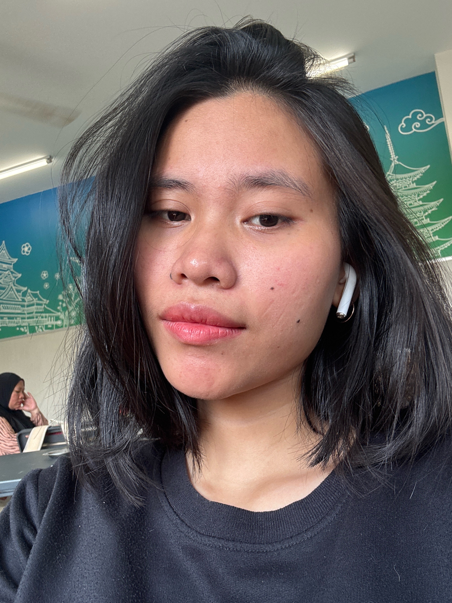 A young woman with medium-length dark hair is the subject of a close-up, head-and-shoulders selfie taken indoors. She is wearing a black crewneck sweatshirt and has white wireless earbuds in her right ear. The background features a brightly colored mural with depictions of Asian architecture, specifically a pagoda, and stylized clouds against a green and blue gradient. To the left and slightly out of focus, another person wearing a black hijab and a pink and beige striped top is visible, seated at a table. The environment appears to be a cafe or common area, with fluorescent lighting visible on the ceiling and a hint of other tables and chairs in the background. The overall scene suggests a casual, everyday moment captured in a public indoor space in East Jakarta, Indonesia.