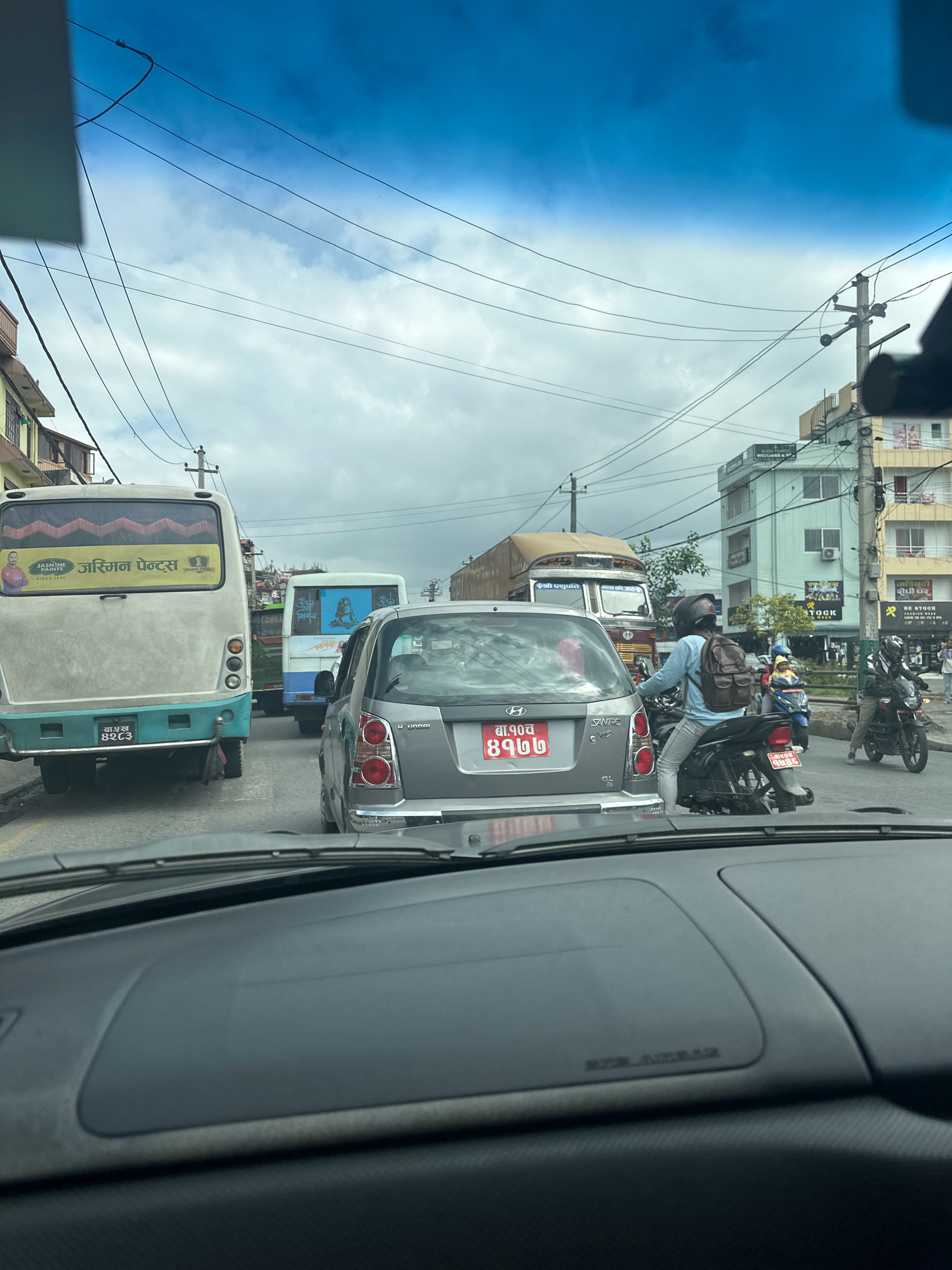 In Kathmandu Metropolitan City, Nepal, a traffic jam is captured from the perspective of a vehicle's dashboard, looking forward through the windshield. A line of vehicles, including a light-colored bus with "Jasmine Paints" branding, a grey sedan with Nepali license plates, and multiple motorcycles, are stationary on a paved road. The bus displays a mural of a man and text in Nepali. A truck with Nepali text is visible further ahead. Buildings line the street, with power lines overhead. The sky is overcast with grey clouds.