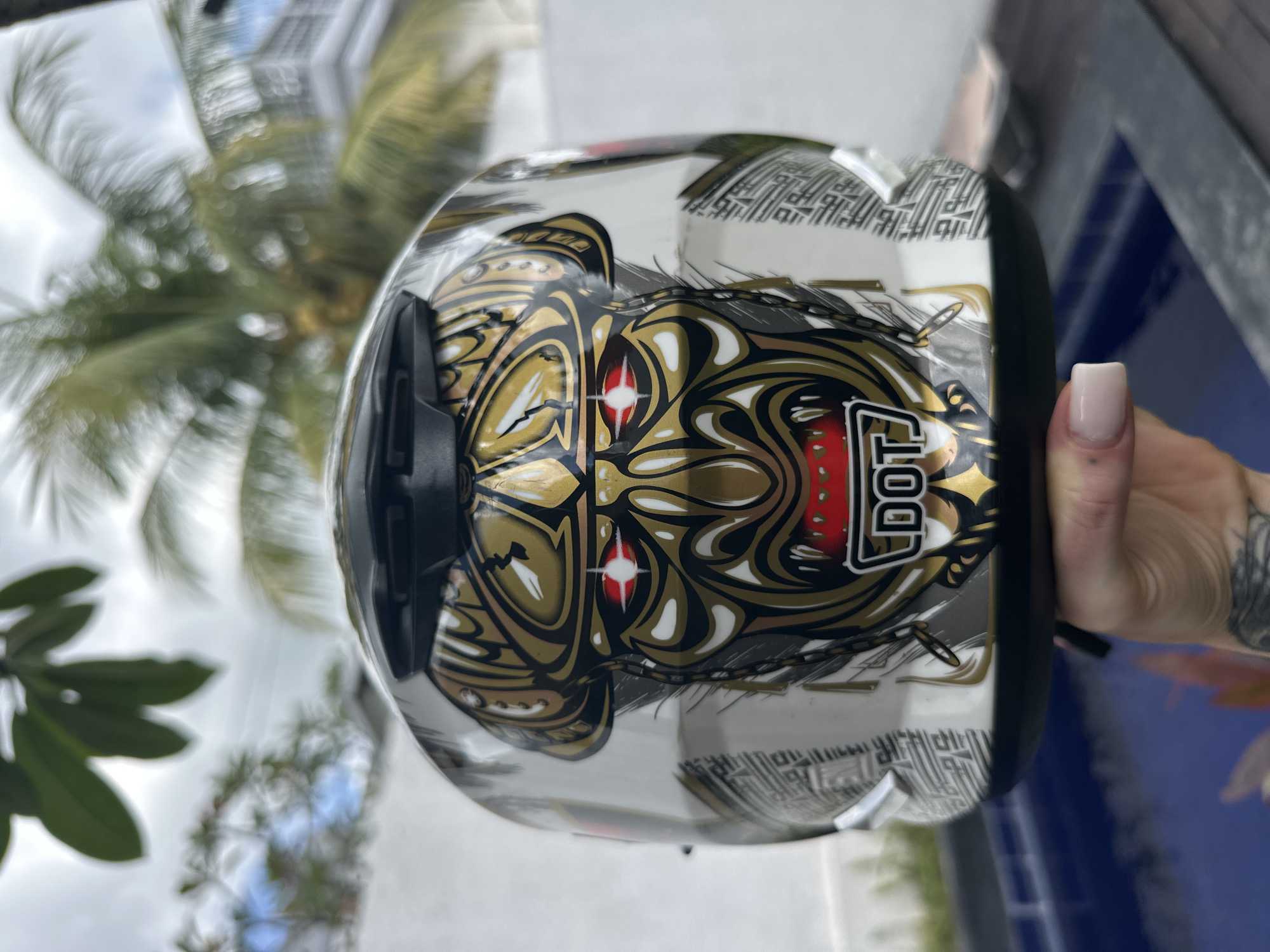 The image shows a motorcycle helmet held by a person with tattooed arm, positioned with a palm tree in the blurred background. The helmet has a white base with a gold and black design featuring a stylized face. The DOT label is visible on the helmet. The scene appears to be outdoors, potentially near a pool or water feature, based on the blue tile-like surface visible in the background. The location is Dalung, Indonesia.
