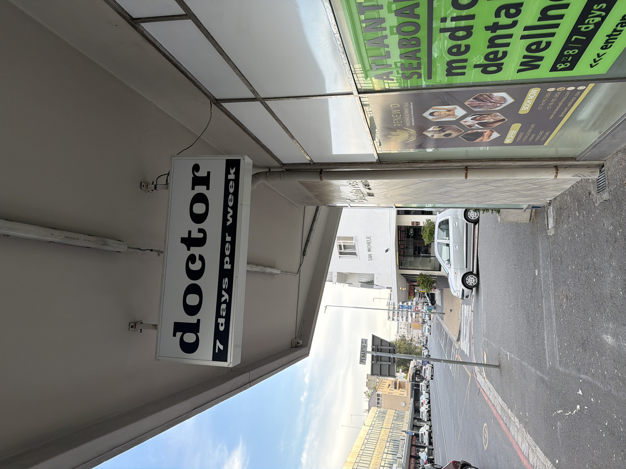 The image shows a street scene in Cape Town, South Africa. A sign for a "doctor" open "7 days per week" is visible under an awning. Next to it, another sign advertises "Atlantis Seaboard" for "medical, dental, wellness," and "Renew'd Wellness & Medi Spa." Across the street, a building labeled "San Michele" is visible. A street sign reading "Penarth" is also visible. Cars are parked along the street. A silver car is parked close to the curb. The overall impression is of a busy urban environment with medical and wellness-related businesses.