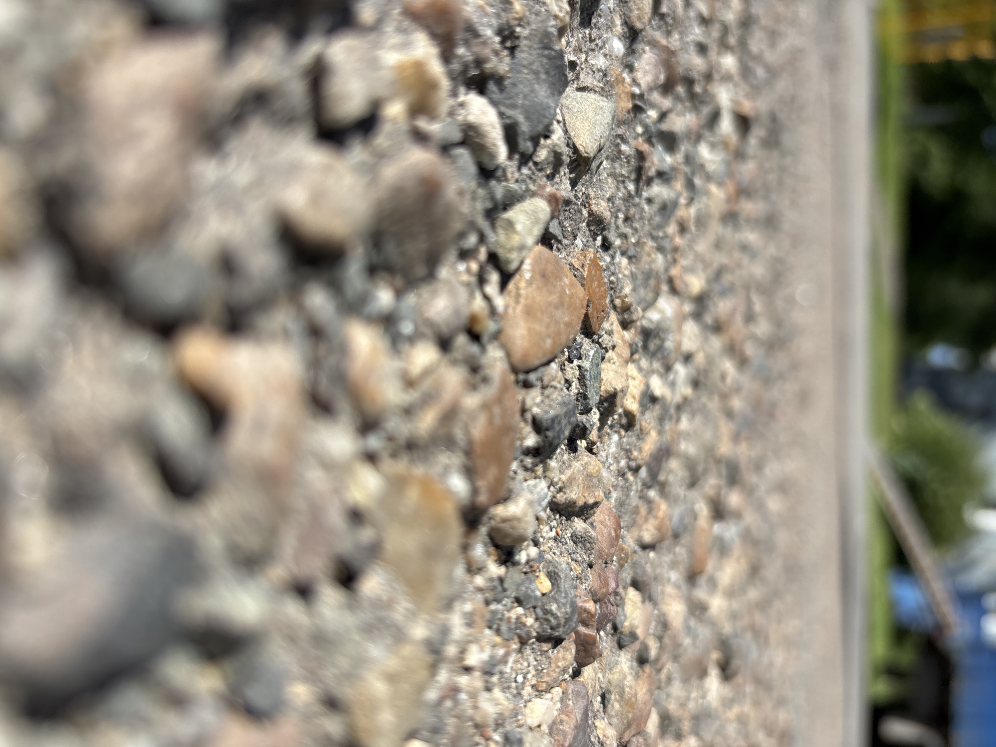 The image shows a close-up, macro view of a textured concrete surface, likely an outdoor wall or pathway. The concrete is composed of various aggregate sizes, from fine grains to larger pebbles and stones, ranging in color from light gray and beige to darker grays and browns. Some stones show smoother, rounded surfaces, while others are more angular.
The depth of field is shallow, with the foreground elements being blurred and the midground sharply in focus. To the right of the concrete, a lighter colored vertical surface is visible, possibly the edge of another structure or a painted line. Beyond this, the background is heavily blurred, showing hints of green foliage and what appears to be man-made objects, possibly a blue bin or container. The lighting suggests it is daytime, with bright sunlight creating highlights on the textured surface of the concrete. There are no people or animals visible in the frame, and no text is discernible. The overall impression is one of an ordinary, everyday outdoor scene, focusing on the tactile and visual details of the concrete's composition.