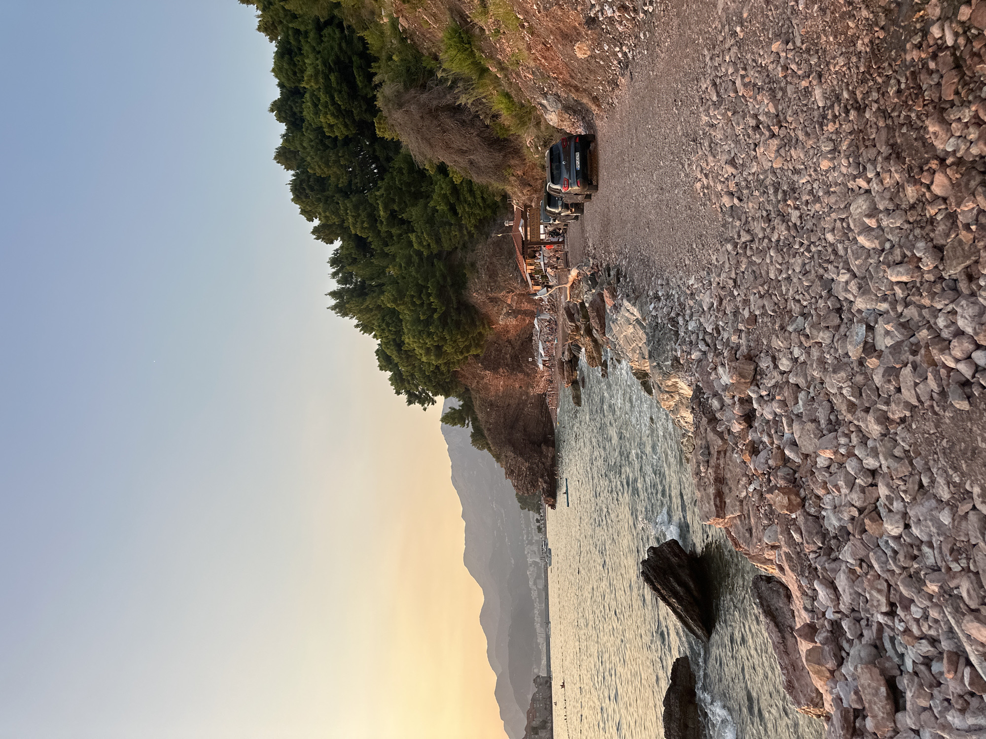 The image captures a scenic view of a coastal road in Sveti Stefan, Montenegro, during what appears to be sunset. The road is unpaved and covered with gravel and rocks, winding along the coastline. On the left side of the road, a rocky shore meets the calm, slightly wavy sea, with a few large rocks visible in the water. Beyond the sea, a town with buildings is nestled against a backdrop of distant, hazy mountains.
On the right side of the road, lush green trees cover a steep, rocky incline. In the mid-ground, a small restaurant or beach bar with an outdoor seating area is visible, with umbrellas providing shade. Several people are seen seated at the establishment and on the beach area to its left.
A line of vehicles, including three dark-colored SUVs and one dark SUV, are parked on the gravel road, facing away from the viewer. The sky is a gradient of soft yellow and blue, indicative of a sunset or sunrise. The overall impression is one of a tranquil coastal setting with a touch of activity from the nearby establishment and parked cars.