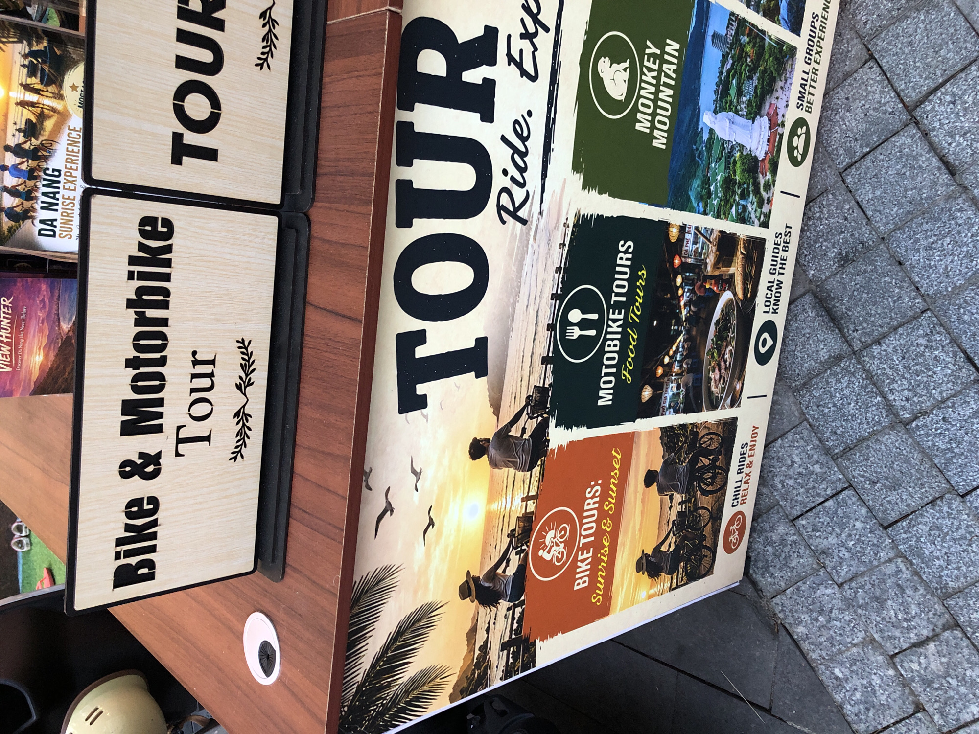 The image depicts a tourist information kiosk, likely located in Đà Nẵng, Vietnam. Two wooden signs with black stencil lettering stand on the kiosk. The left sign reads "Bike & Motorbike Tour," and the right sign reads "TOUR." Below these signs, a large, colorful poster advertises various tours. The poster features three distinct sections. The left section, with an orange background, highlights "BIKE TOURS: Sunrise & Sunset." It shows a silhouette of two people riding bicycles along a beach at sunset, with seagulls flying overhead. The middle section, with a dark background, advertises "MOTORBIKE TOURS: Food Tours," and displays images of local Vietnamese cuisine. The right section, with a green background, promotes "MONKEY MOUNTAIN," and shows a view of the Son Tra Peninsula with a large white statue.
Below the advertised tours on the poster, there are icons and text related to tour features. The "BIKE TOURS" section includes an icon of a bicycle and text that says "CHILL RIDES RELAX & ENJOY." The "MOTORBIKE TOURS" section has a location pin icon and text reading "LOCAL GUIDES KNOW THE BEST." The "MONKEY MOUNTAIN" section features an icon of people and text that says "SMALL GROUPS BETTER EXPERIENCING."
The kiosk itself is made of dark wood, and the surrounding ground is paved with grey interlocking tiles. In the background, other promotional materials and what appears to be part of a street or walkway are visible. The overall scene suggests an outdoor location where tourists can gather information and book activities.