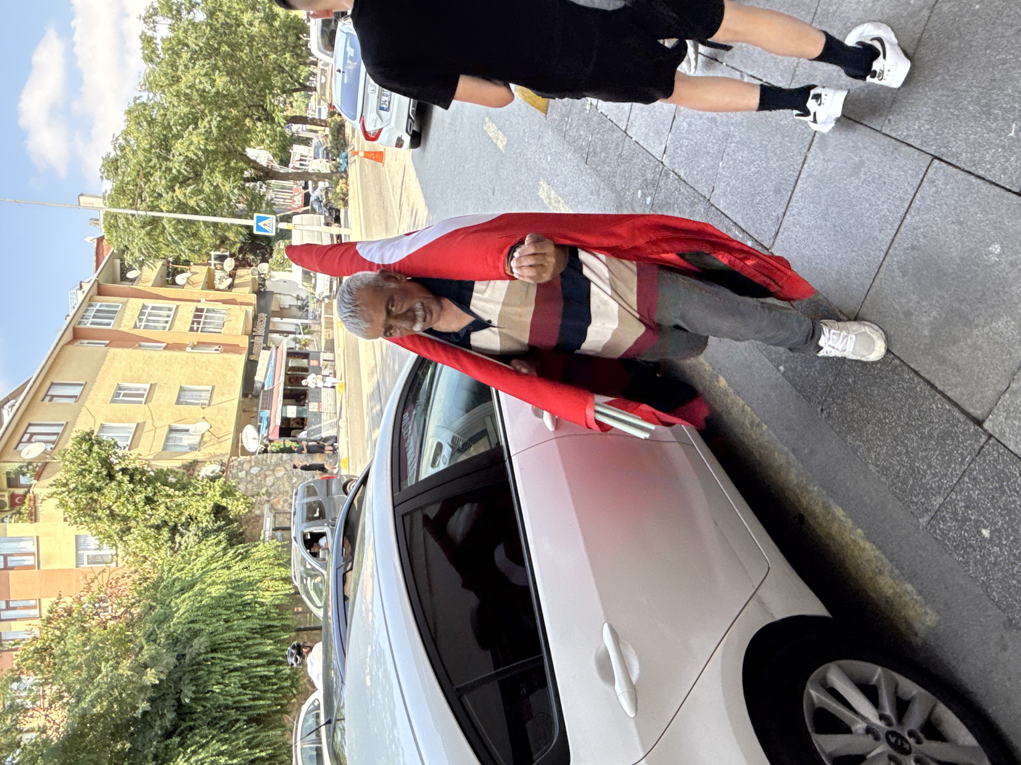 A man, identified as a Turkish supporter of Fenerbahçe S.K., walks along a sidewalk in Kadıköy, Turkey, holding a large Fenerbahçe flag. The flag, predominantly red with a white crescent and star, is draped over his shoulders and held in his right hand. He wears a striped polo shirt and jeans, with a mustache and gray hair. To his right, a younger person in black shorts and a black t-shirt, wearing black socks and white sneakers, walks away from the camera. A white car is parked on the left side of the image. In the background, buildings and trees line the street, with other vehicles and pedestrians visible. The setting appears to be a sunny day on a city street.