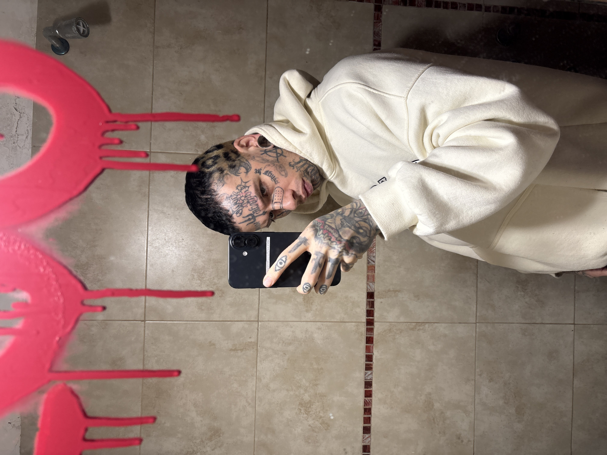 A tattooed person in a light-colored hoodie is taking a selfie in front of a tiled mirror, likely in a bathroom. The mirror is partially obscured by a pink, paint-like design. The environment appears to be a bathroom with neutral-toned tiles on the wall and floor, and a showerhead is visible in the upper left corner. There is no evidence to suggest the location is in Buenos Aires, Argentina.