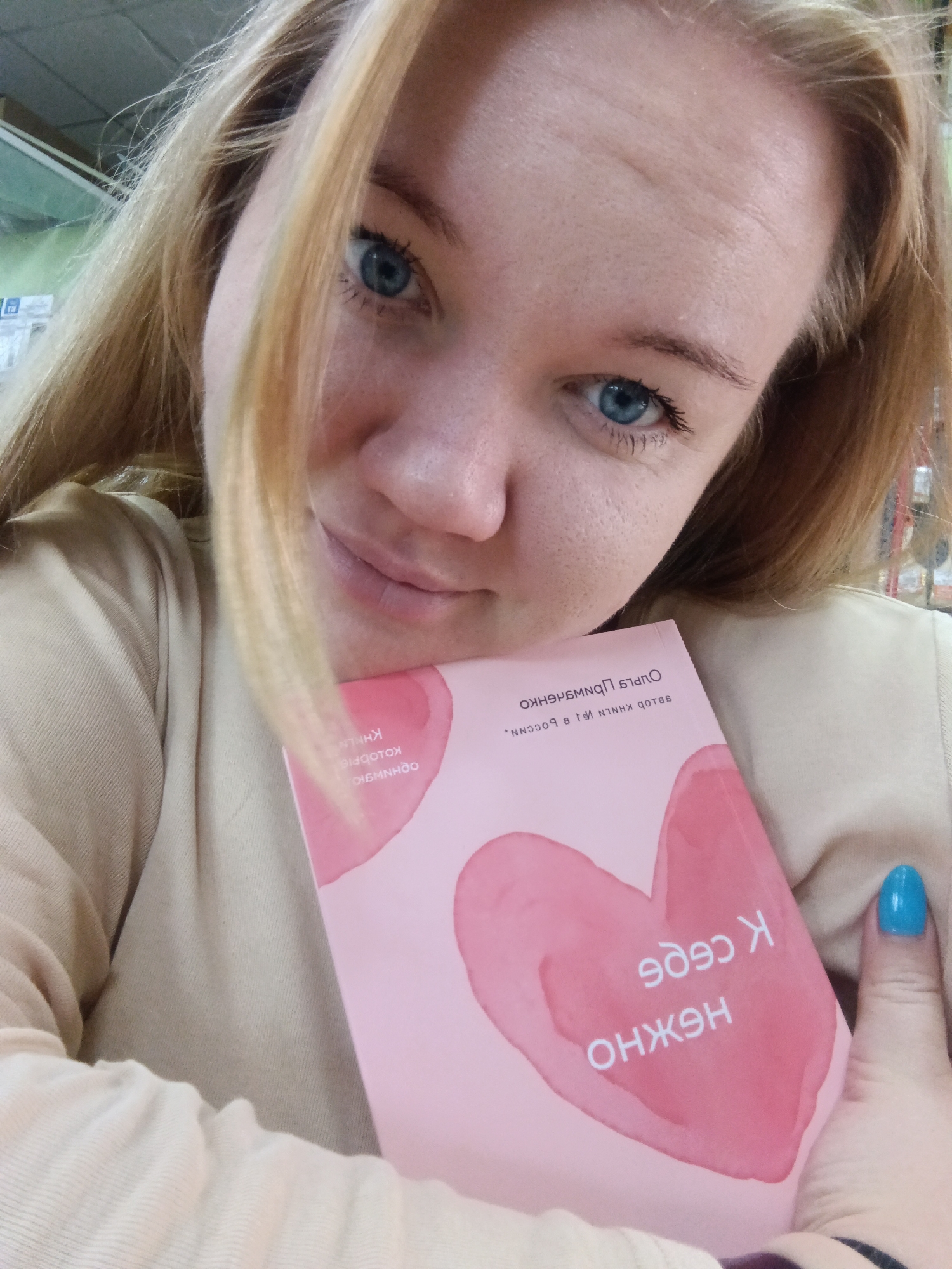 A young woman with blonde hair and blue eyes is taking a selfie in a store. She is holding a pink book with watercolor hearts on the cover, and text in Cyrillic script. Her left hand is visible, with bright blue nail polish. The background appears to be shelves in a retail environment. The text on the book, when deciphered and reversed, appears to read "К себе нежно" (To yourself, tenderly) and "Автор книг для тебя" (Author of books for you). Based on the visual cues and the Cyrillic text, the location could be any retail bookstore in Russia. Without further context or specific landmarks, pinpointing the exact location in the city of Fokino, Russia, is not possible from the image alone.