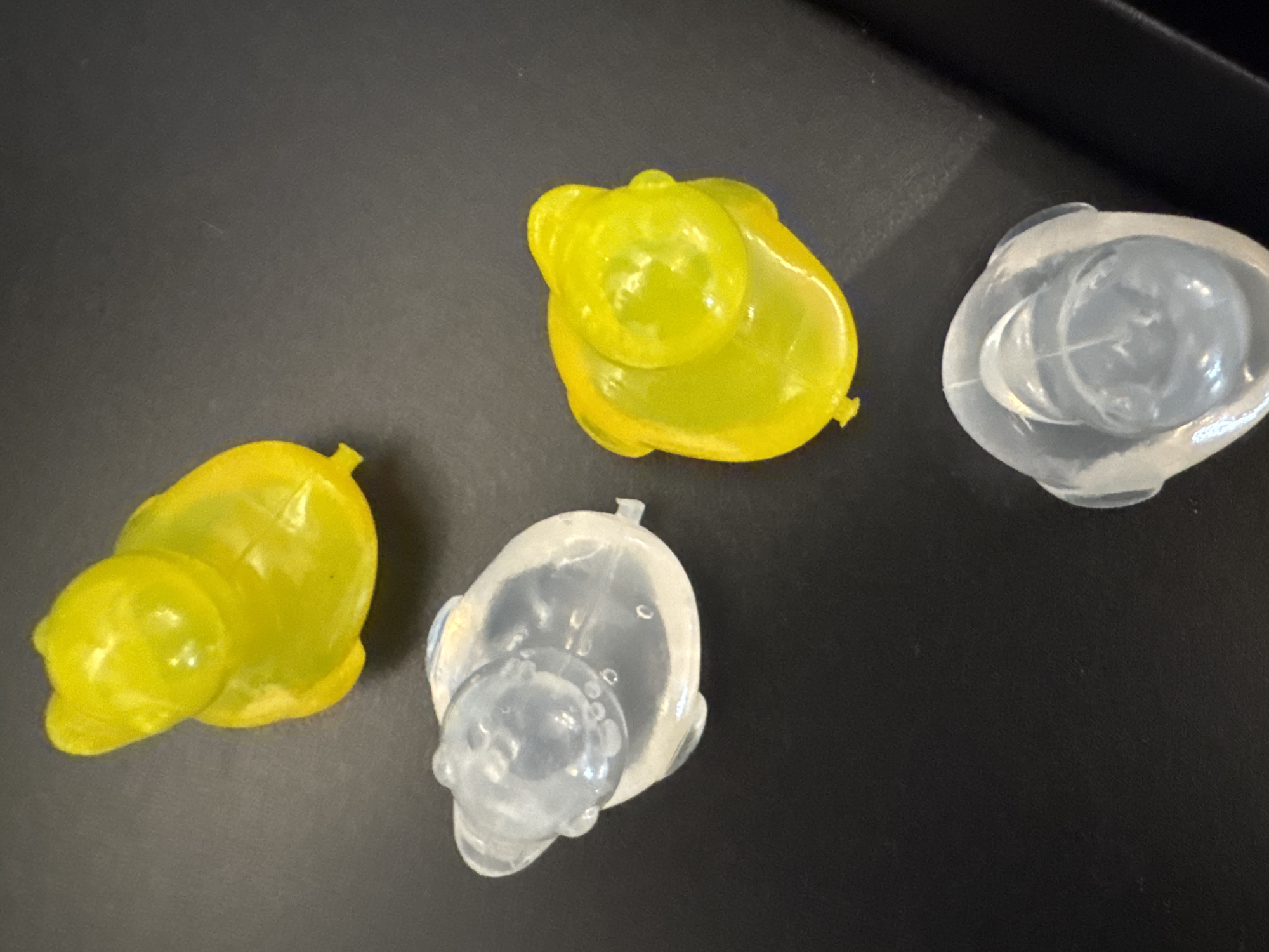 The image shows four small rubber duck toys on a dark, textured surface, possibly a table or countertop. Two of the ducks are bright yellow, and two are translucent white. The ducks are arranged in a somewhat scattered pattern. One yellow duck is in the upper left quadrant, facing downwards and slightly to the left. A translucent white duck is below it and to the left, also facing downwards. Another yellow duck is to the right and slightly below the white duck, facing to the right. A second translucent white duck is at the bottom center of the image, facing upwards. The lighting suggests an indoor setting, but there are no visible windows, doors, or other architectural features to indicate a specific location within Kremenchuk, Ukraine. The surface appears to be a solid material with some faint scuffs or marks.