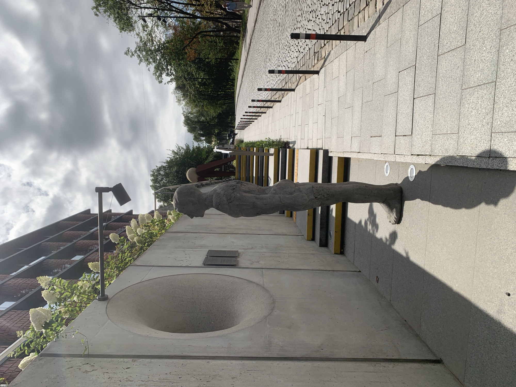 An outdoor scene in Lviv, Ukraine, features a modern building with a brick facade situated behind a concrete wall. White hydrangeas bloom along the top of the wall, cascading down. A concrete sculpture of a standing human figure is positioned in the foreground, facing left, with its shadow cast on the ground. Adjacent to the sculpture are a set of concrete stairs with yellow and grey risers, leading upwards. To the right of the stairs, a cobblestone street is visible, lined with bollards. A man in a light blue shirt and dark trousers walks along the cobblestone street in the background. The sky is overcast with grey clouds.
